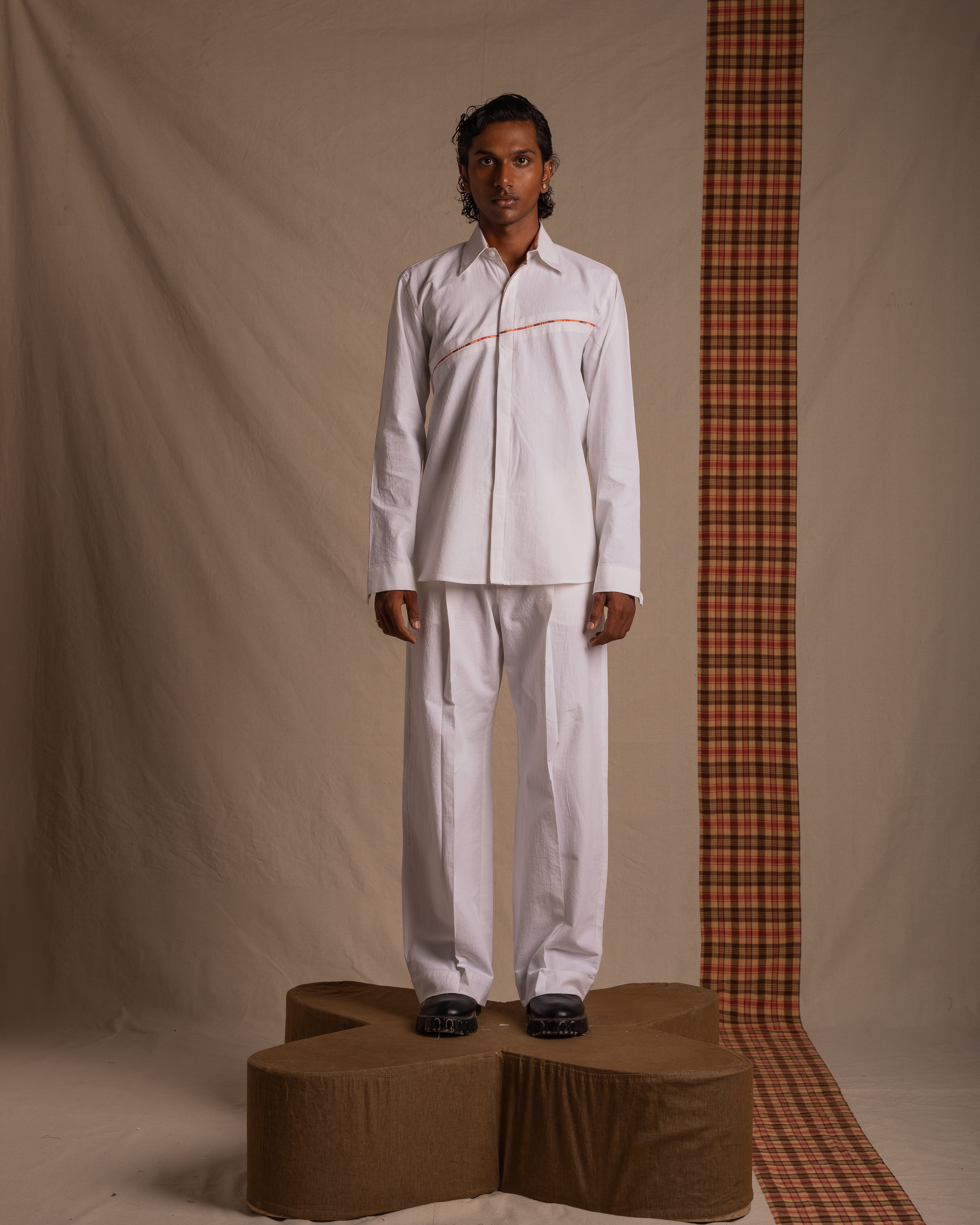 Male model in white button-up shirt and pants with red accent, standing on a brown platform