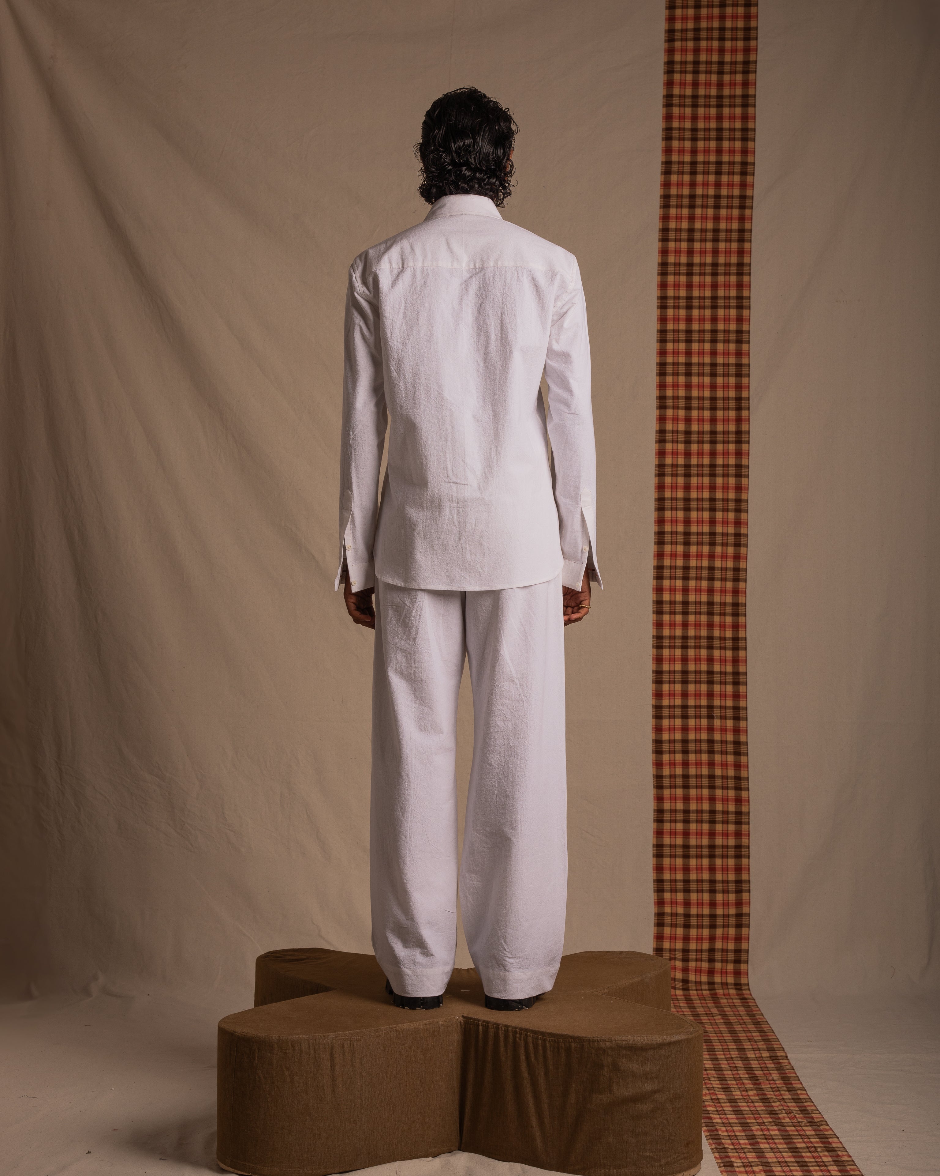 Man in white linen shirt and pants standing on brown platform, minimalist indoor setting