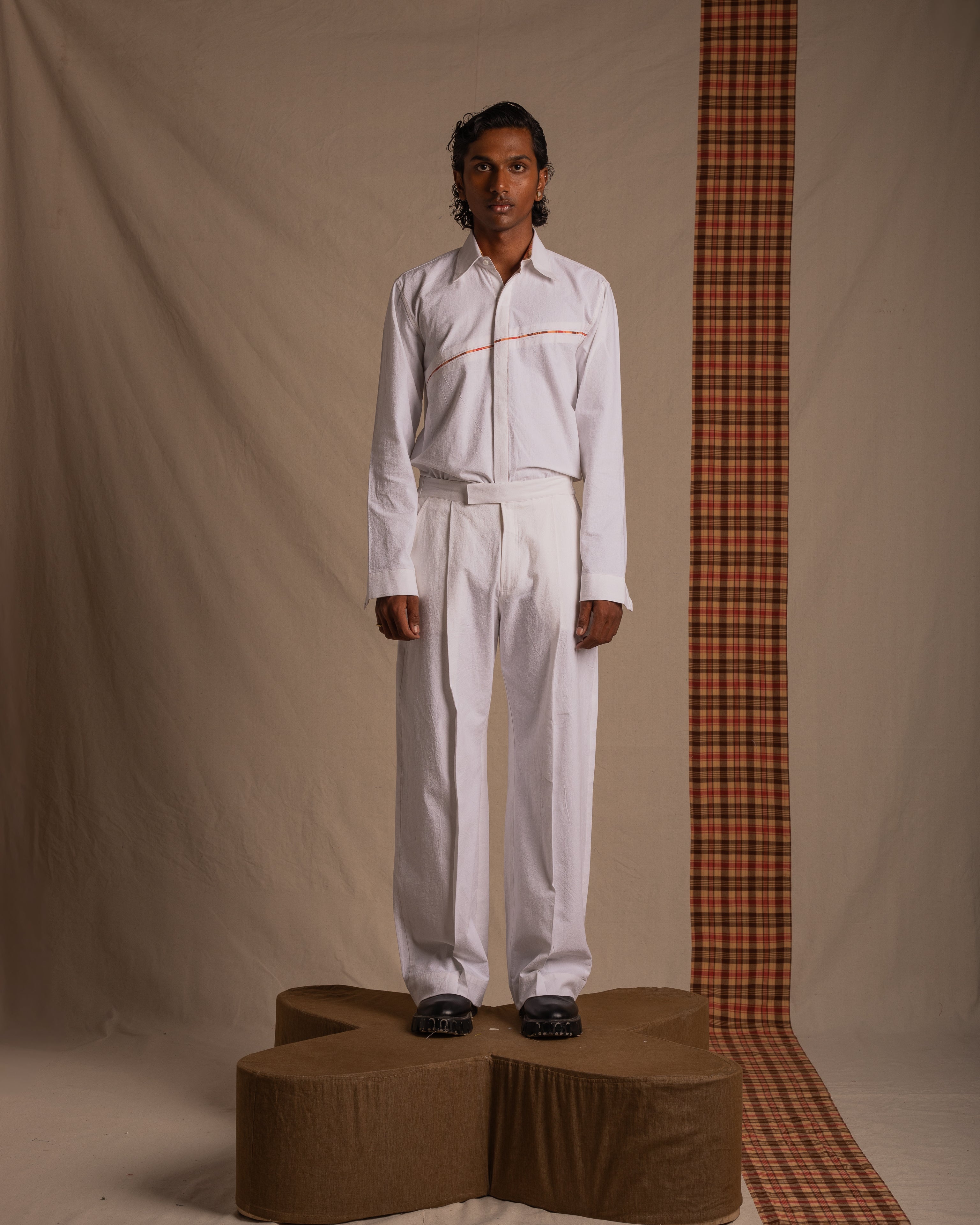 Model in white long sleeve shirt and trousers with black shoes, standing on a brown platform against a beige background with a plaid fabric accent, streetwear fashion