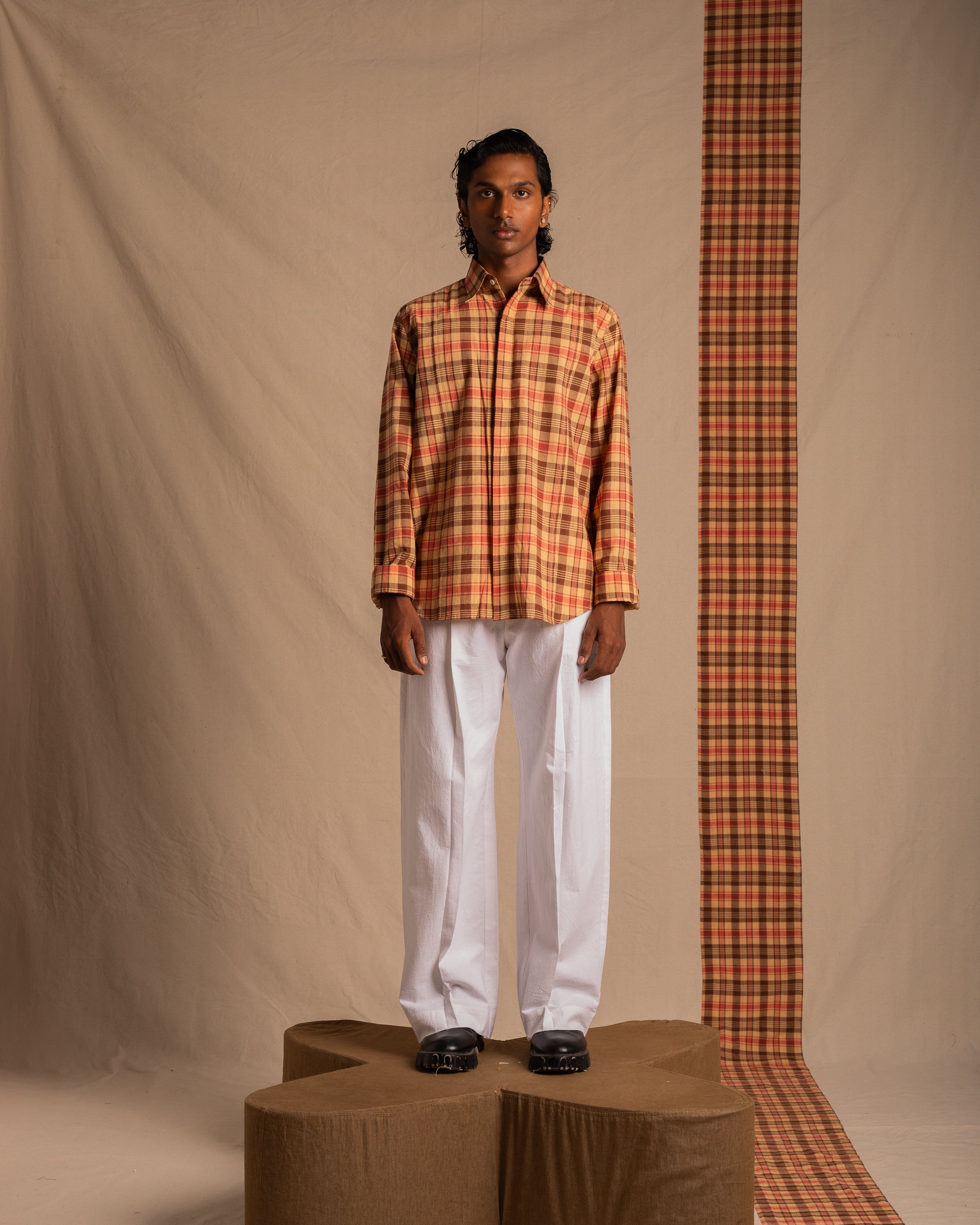 Male model wearing a plaid button-up shirt with white pants and black shoes, standing in a studio with beige and plaid backdrop, streetwear fashion look.