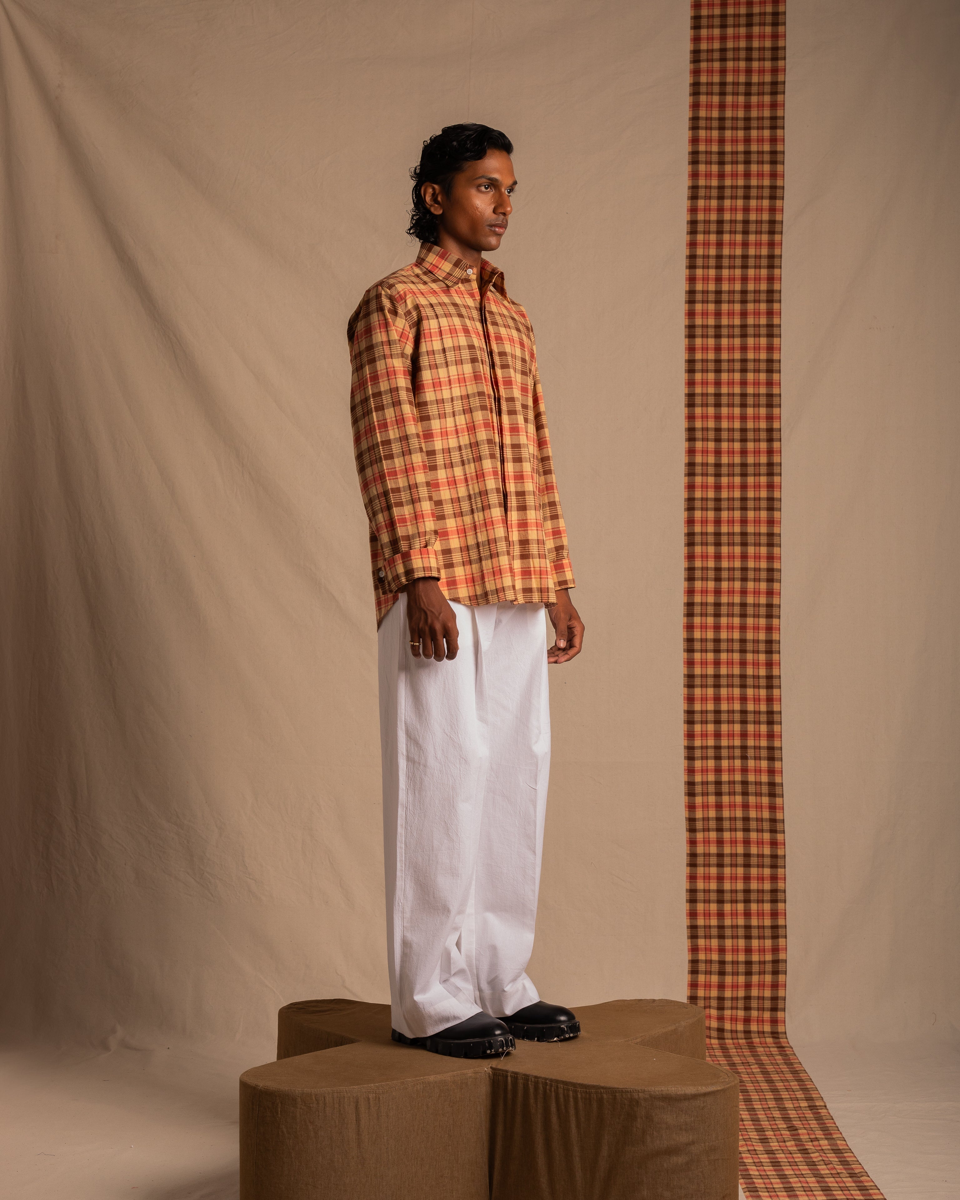 Man in orange plaid shirt and white wide-leg pants standing in minimalist studio