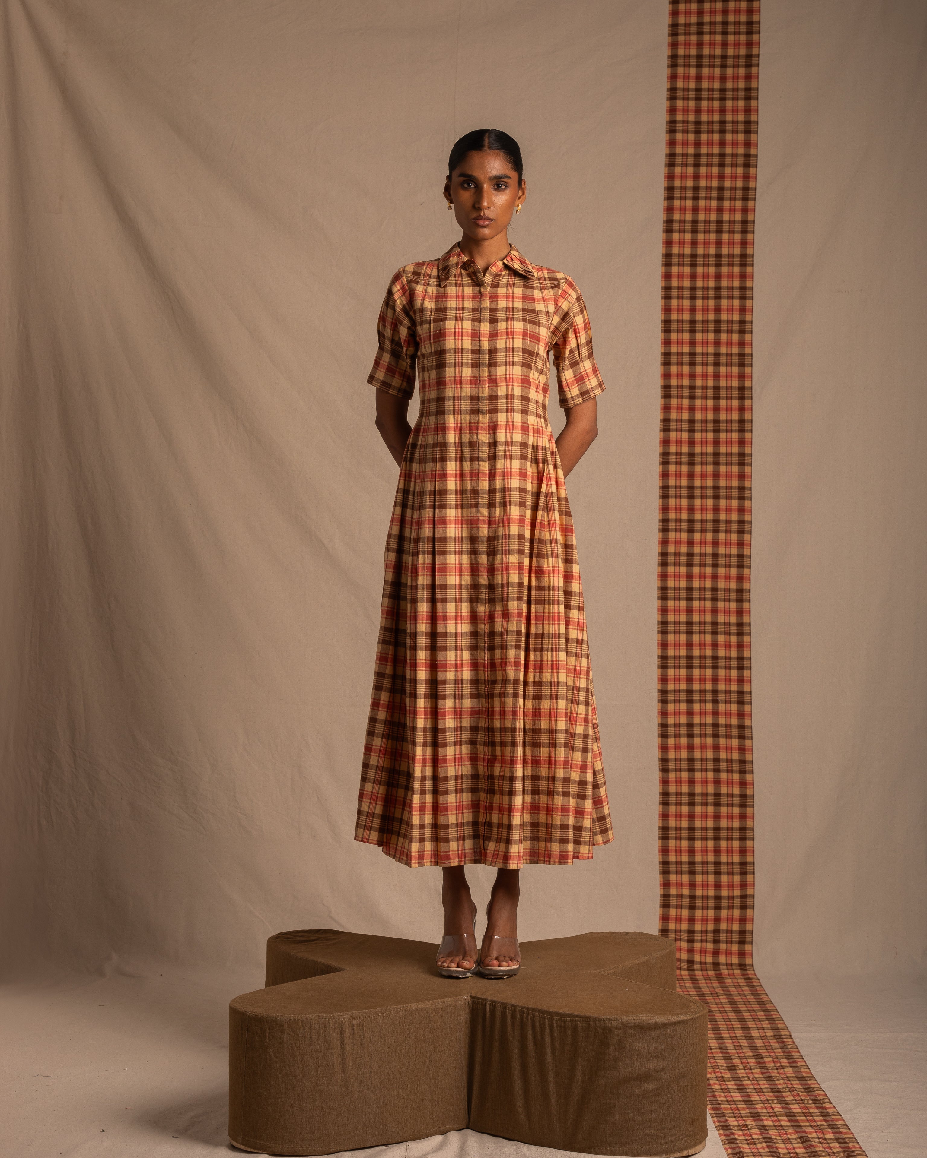 Woman in a brown and red plaid shirt dress, standing on a geometric platform in a studio setting