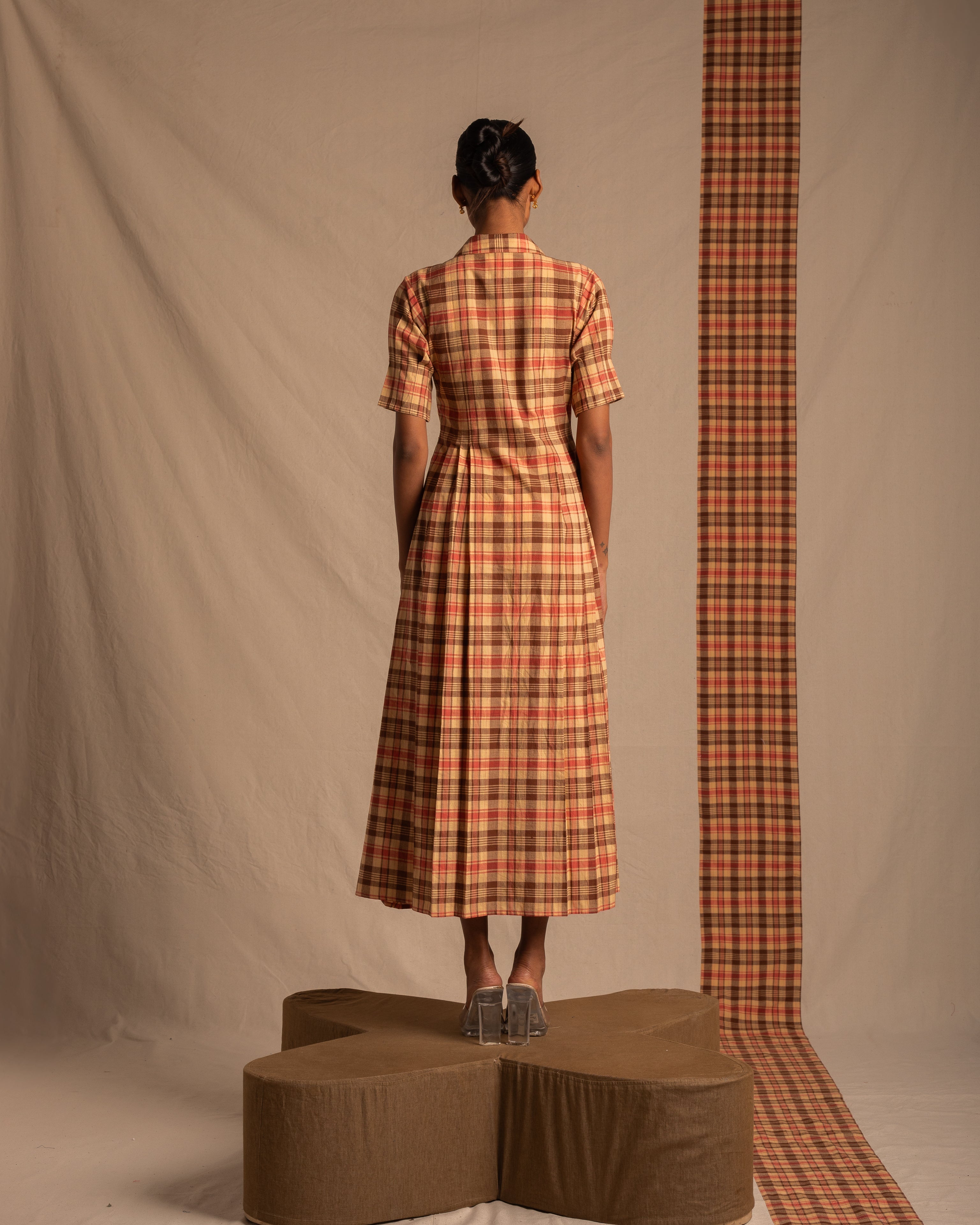Woman in a plaid midi dress with short sleeves, standing on a brown platform, viewed from behind.
