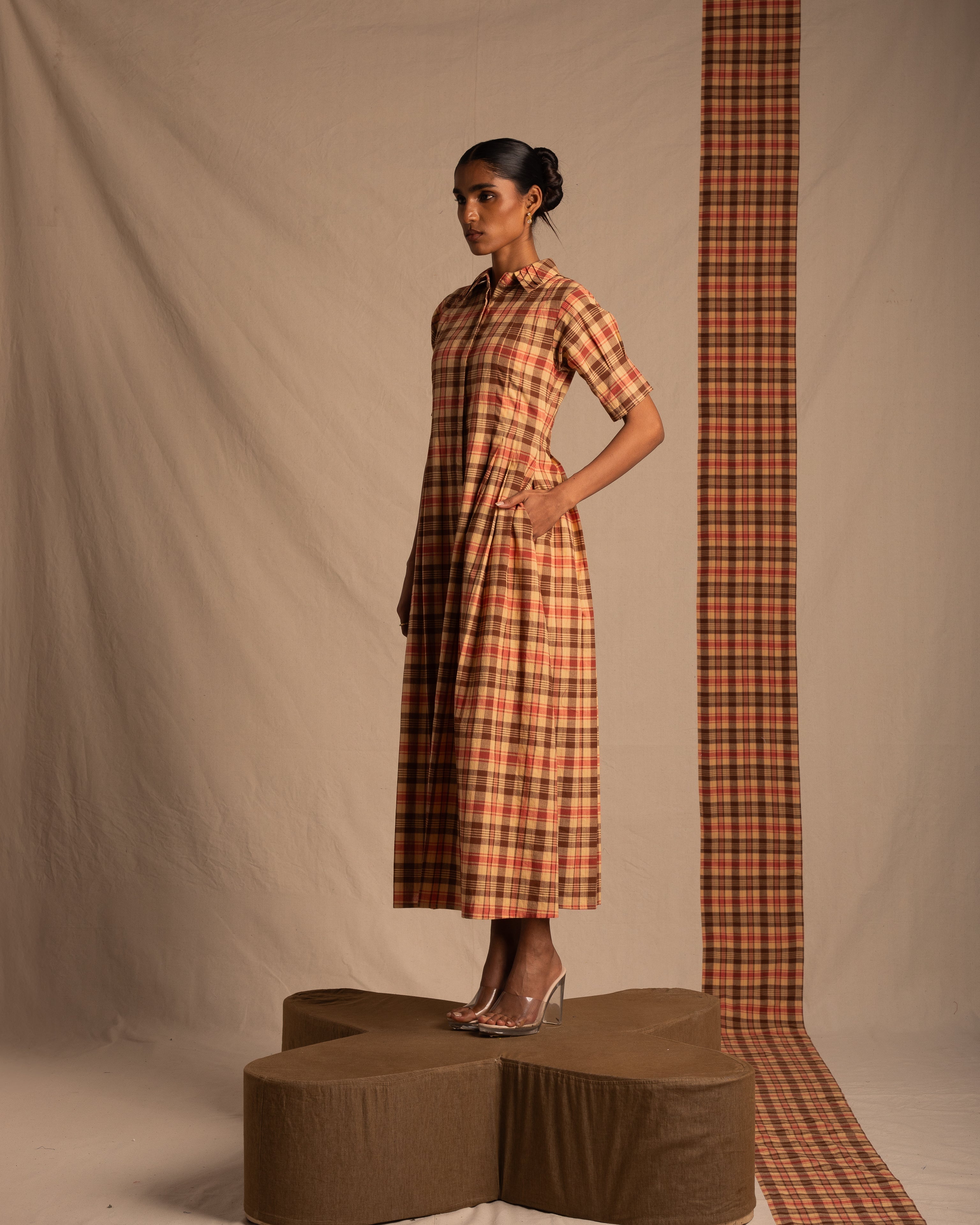 Woman in plaid midi dress with short sleeves, clear heels, standing on geometric platform against neutral backdrop.