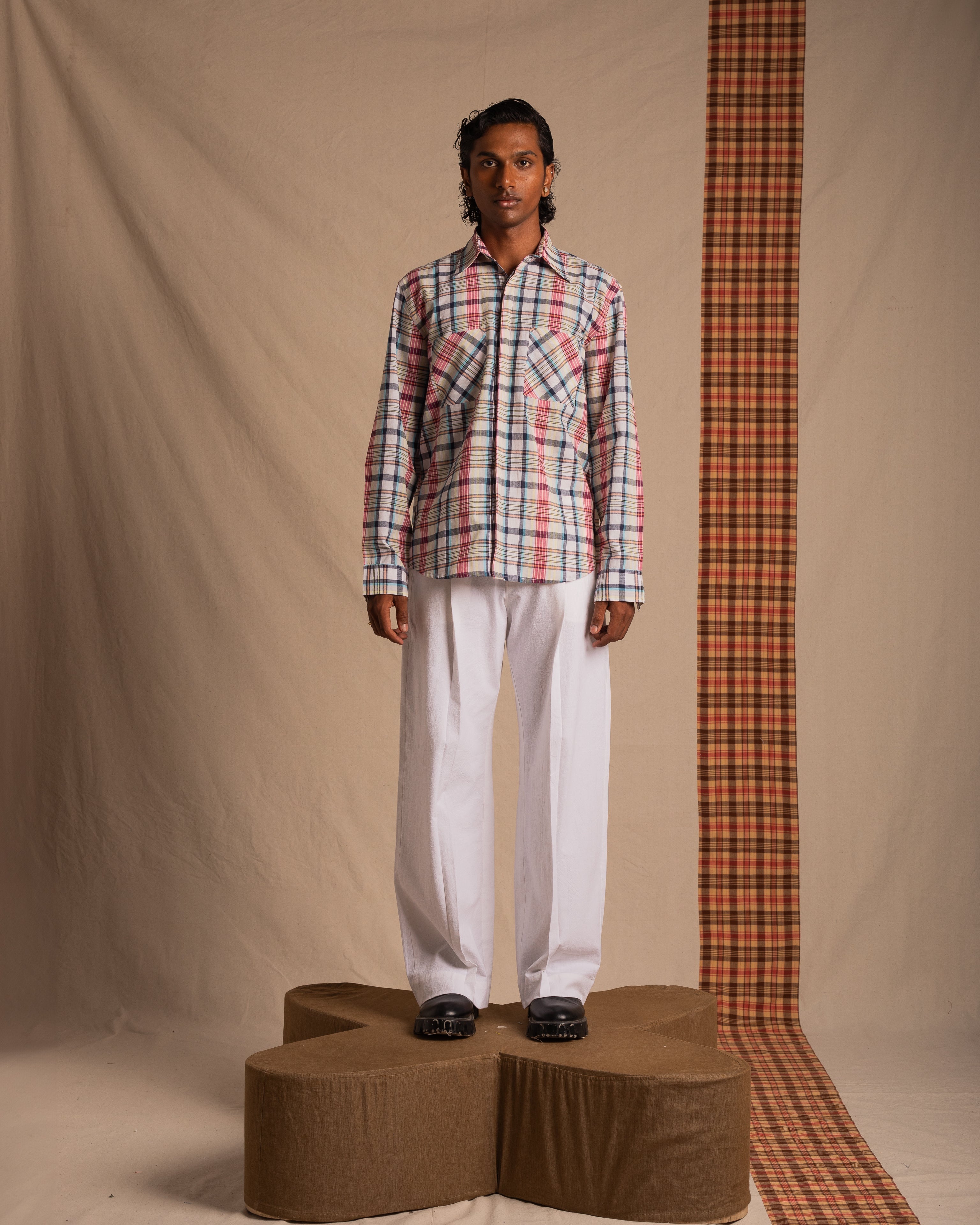Man wearing a plaid shirt with white pants and black shoes, standing on a platform in a studio setting.