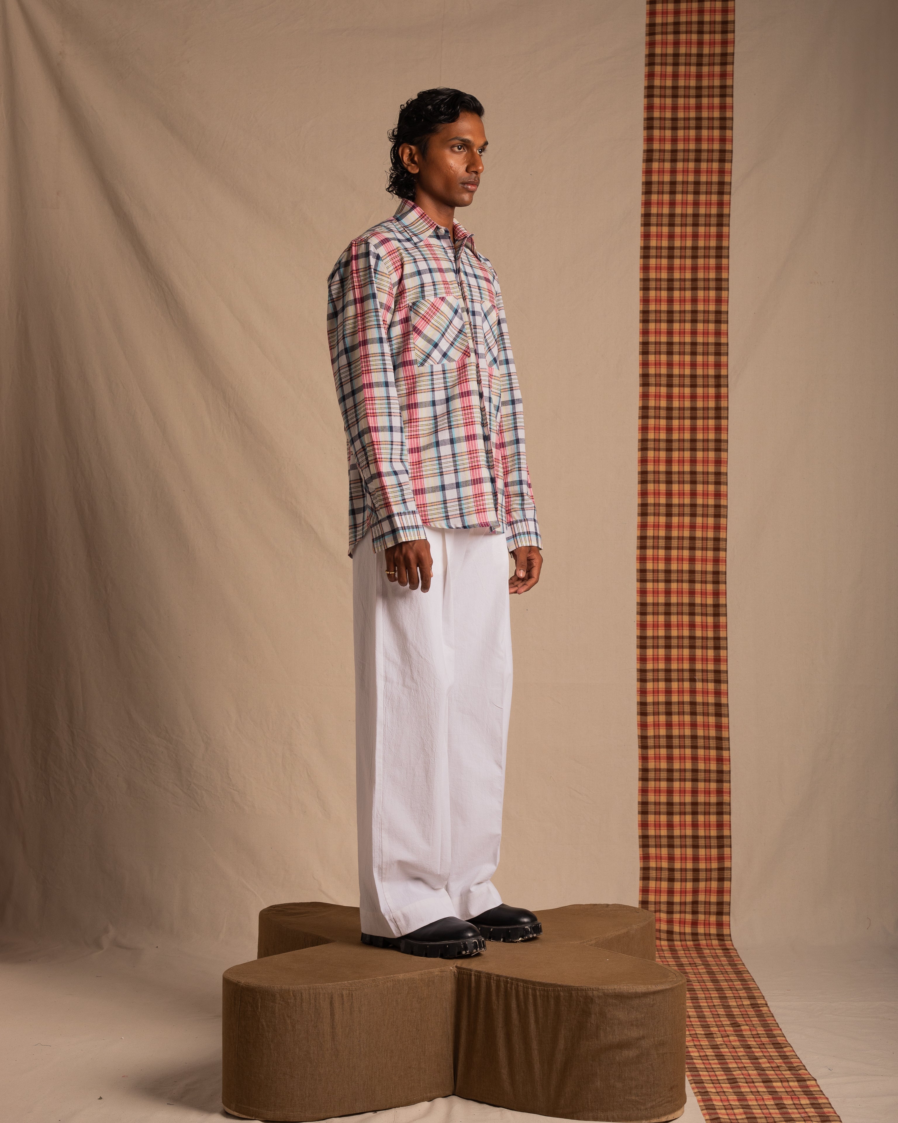 Man wearing plaid shirt, white wide-leg pants, and black shoes on beige studio set