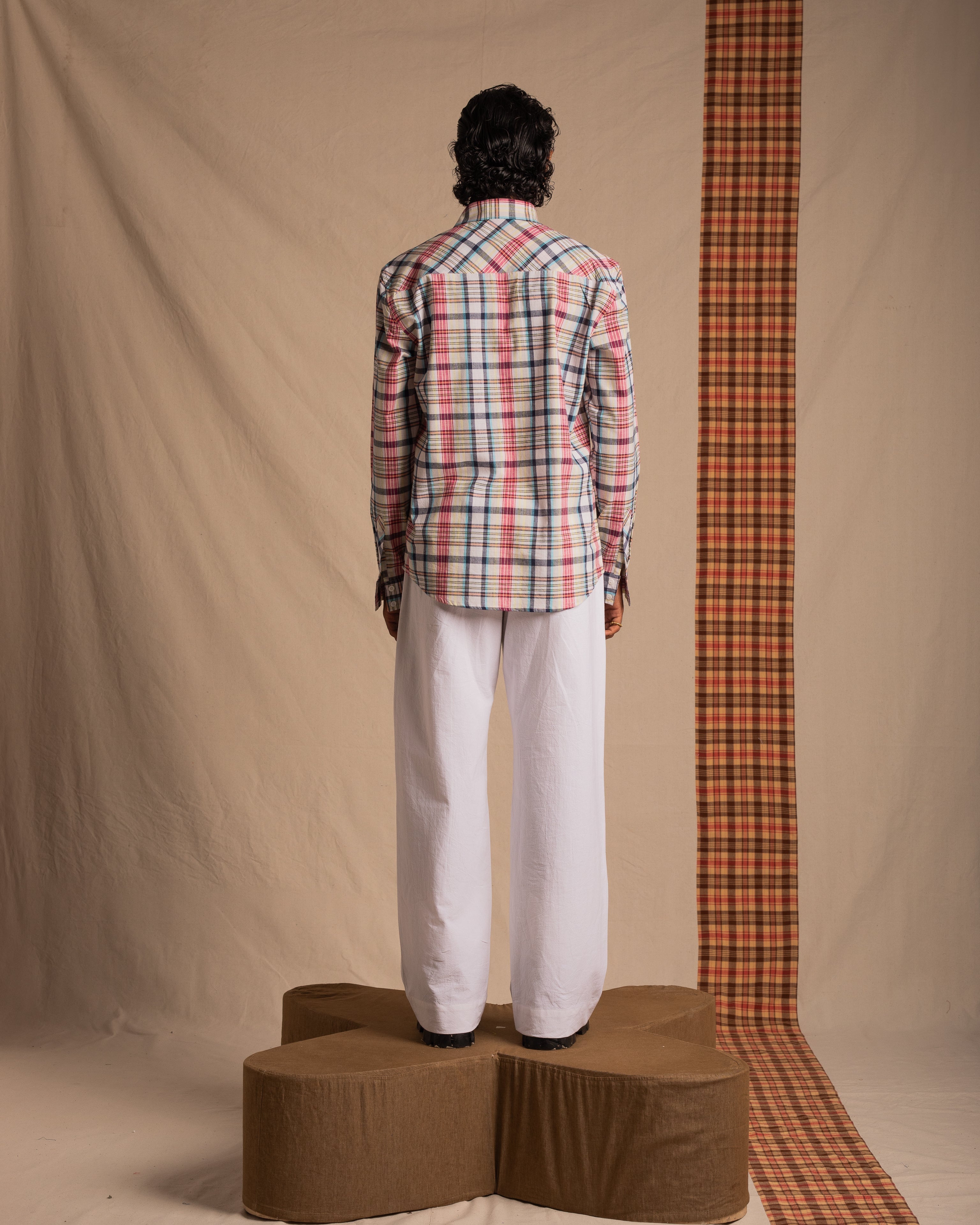 Man in plaid button-up shirt and white pants standing on brown platform, back view
