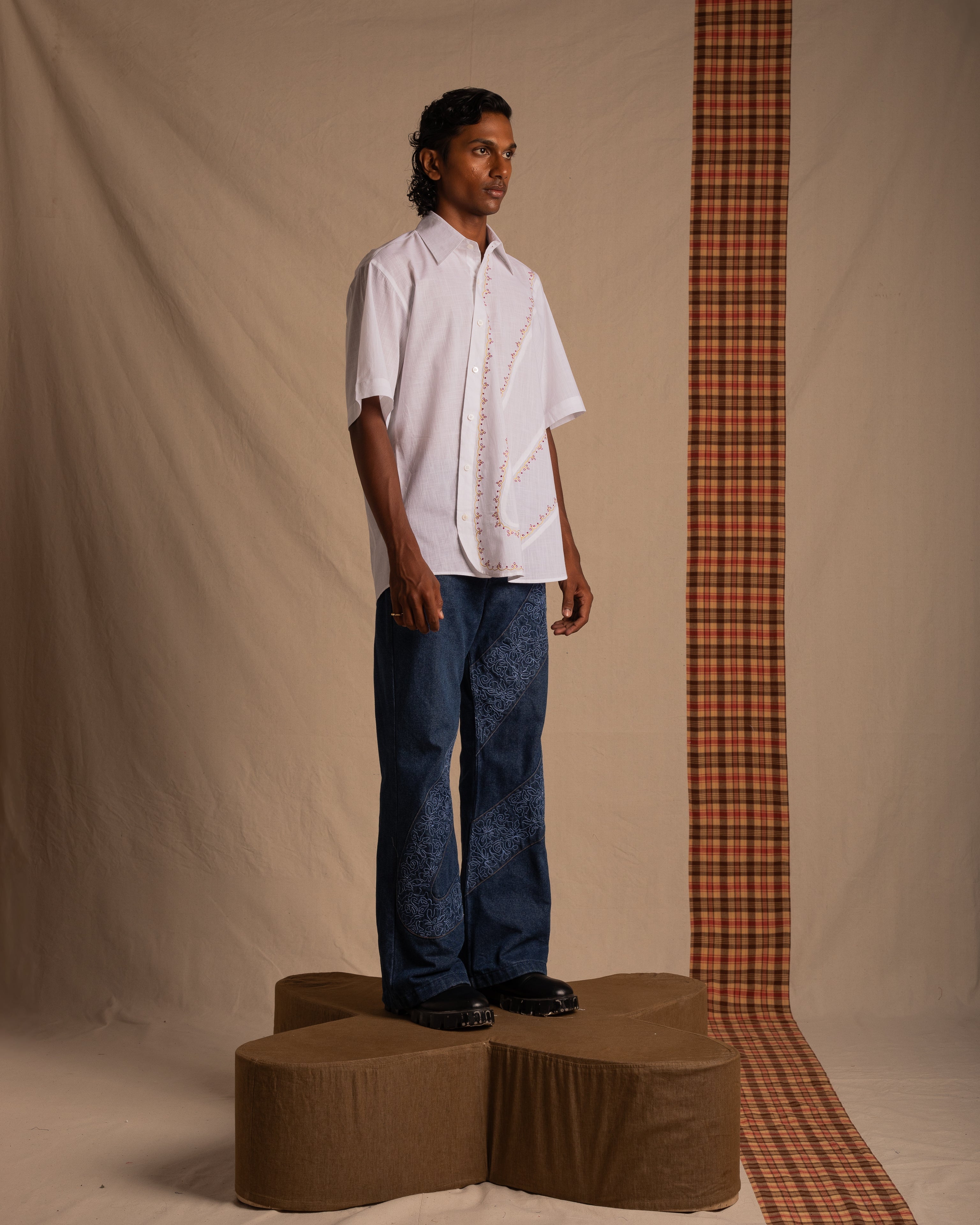 Man modeling streetwear in white short-sleeve shirt, blue pattern jeans, chunky shoes indoors