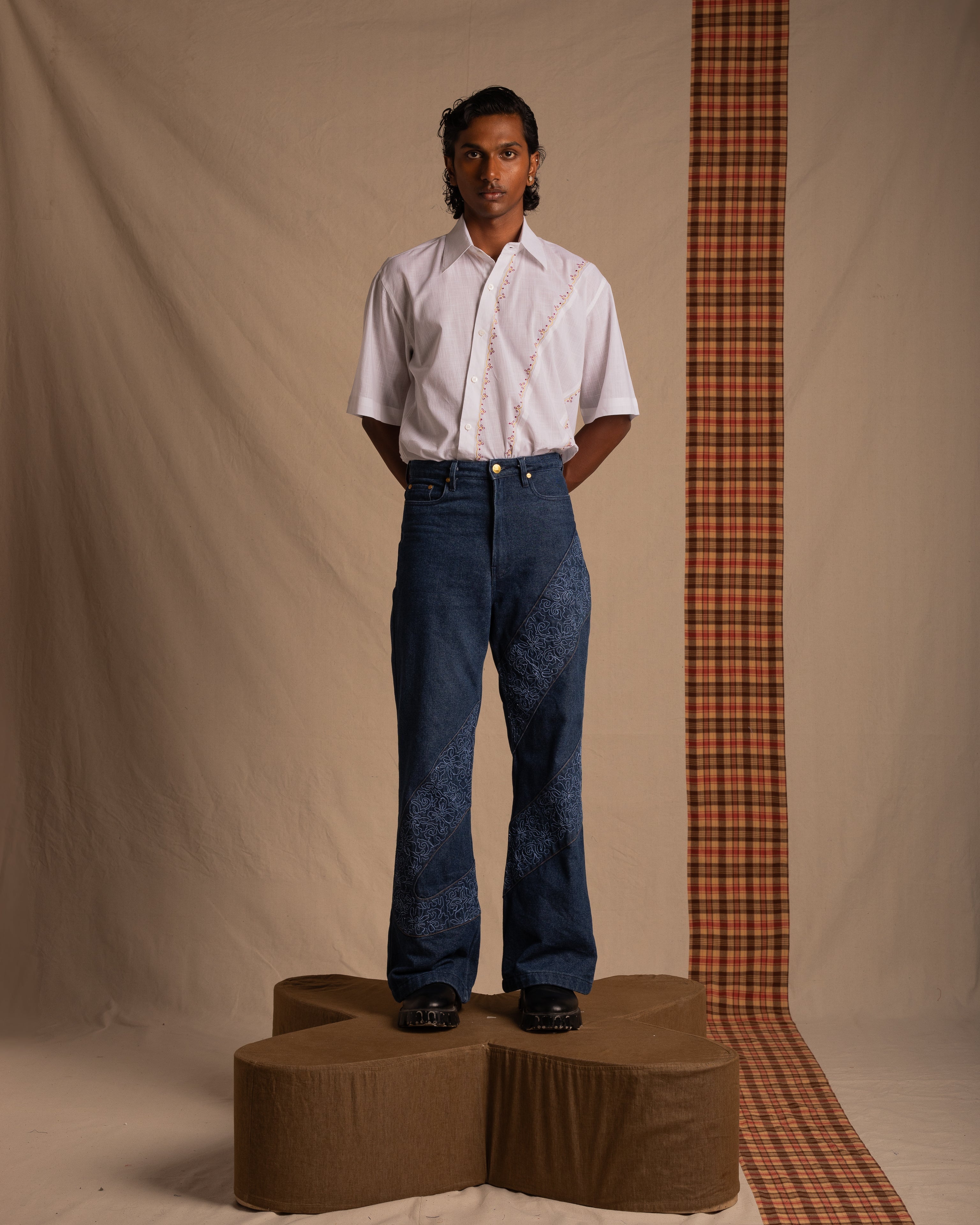 Man modeling streetwear with a white short sleeve shirt and blue embroidered jeans, standing on a geometric platform in a minimalist studio.