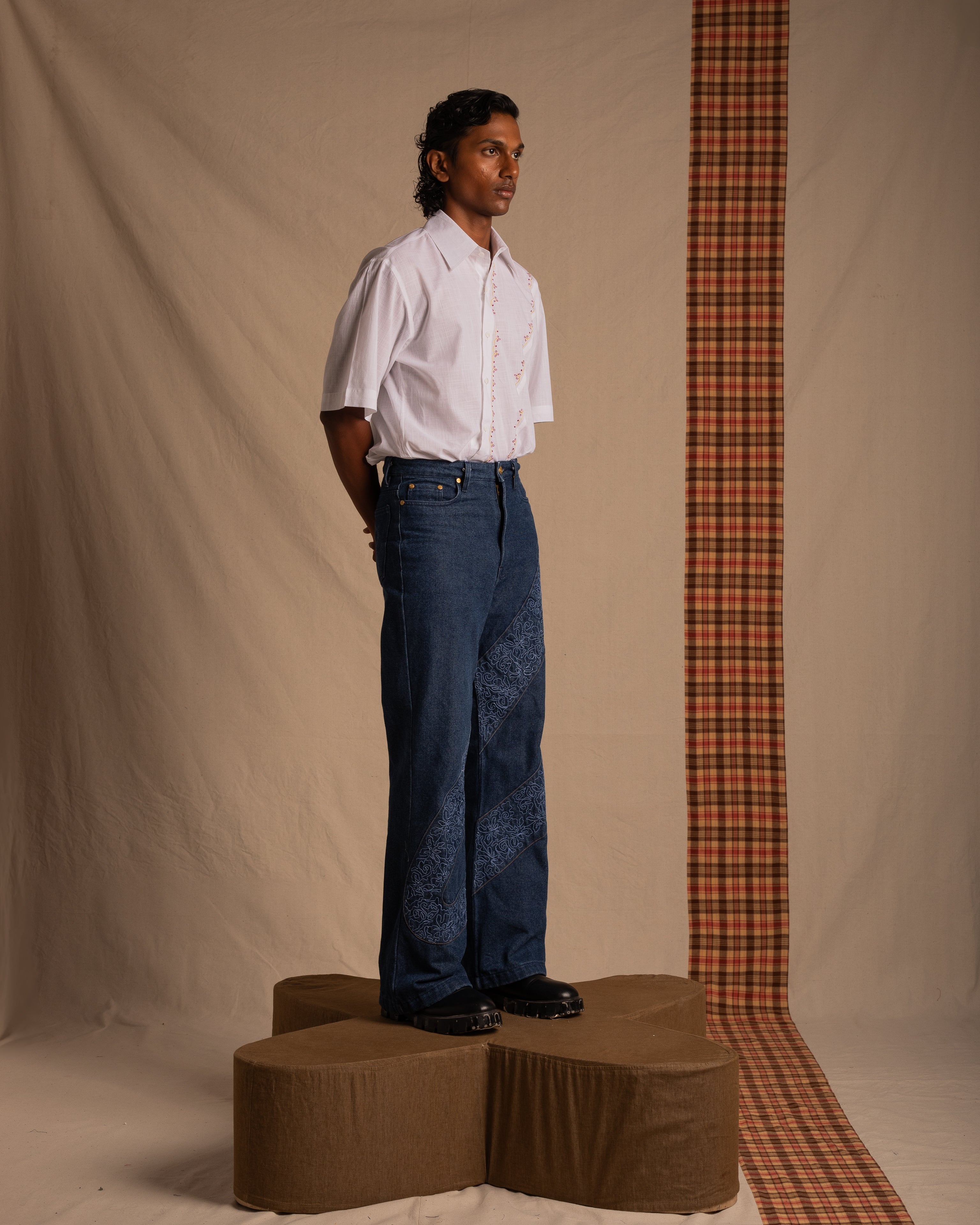 Model in white short sleeve shirt and blue patterned wide-leg jeans, standing on brown platform with plaid fabric backdrop, streetwear fashion