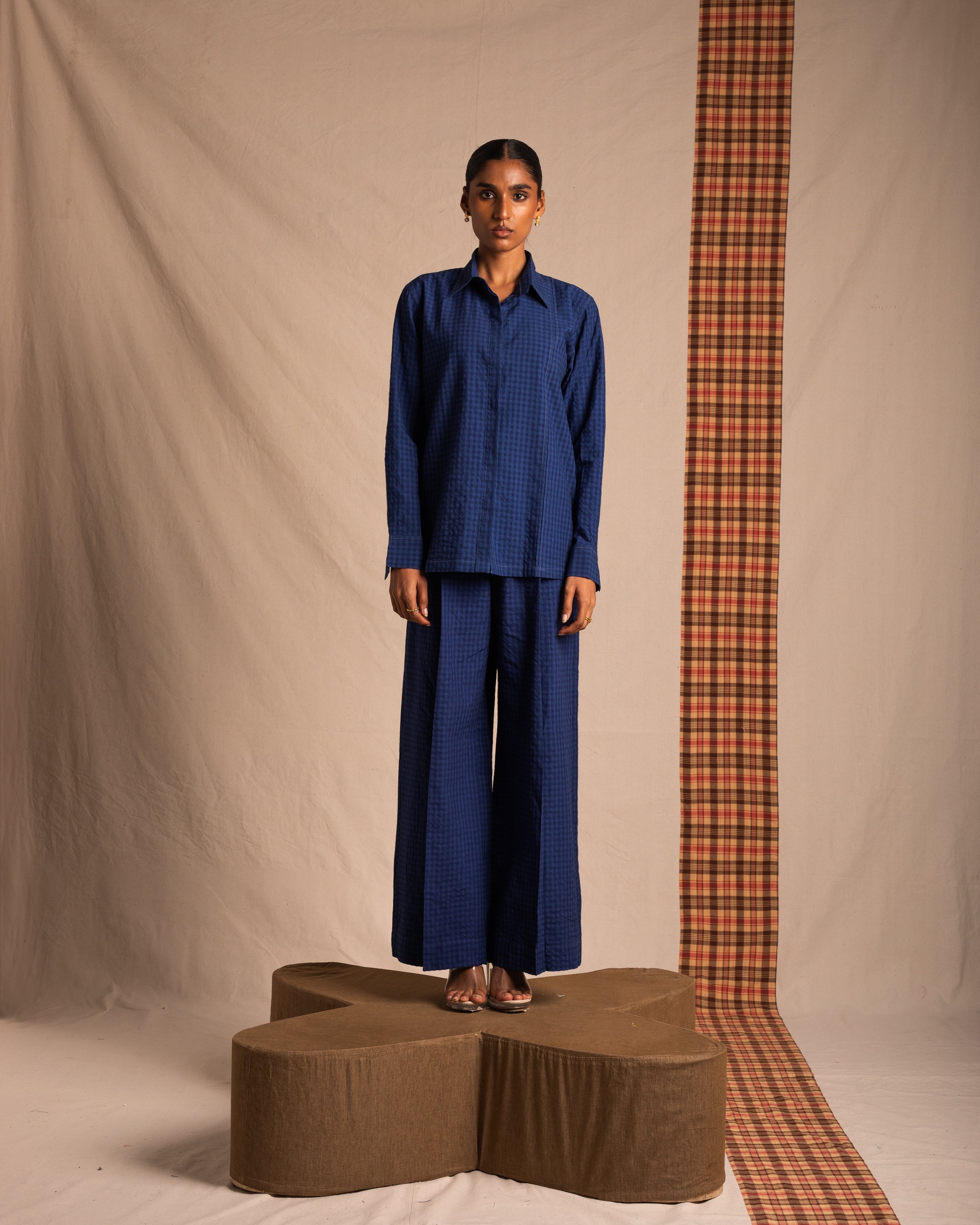 Woman wearing blue checkered long-sleeve shirt and wide-leg pants, standing on brown platform