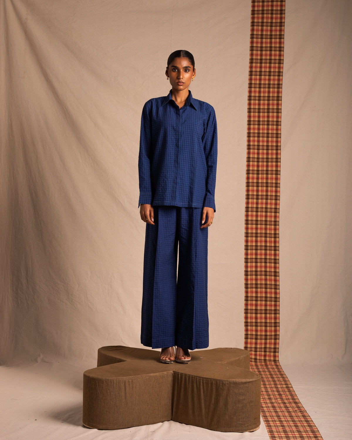 Woman wearing blue checkered long-sleeve shirt and wide-leg pants, standing on brown platform