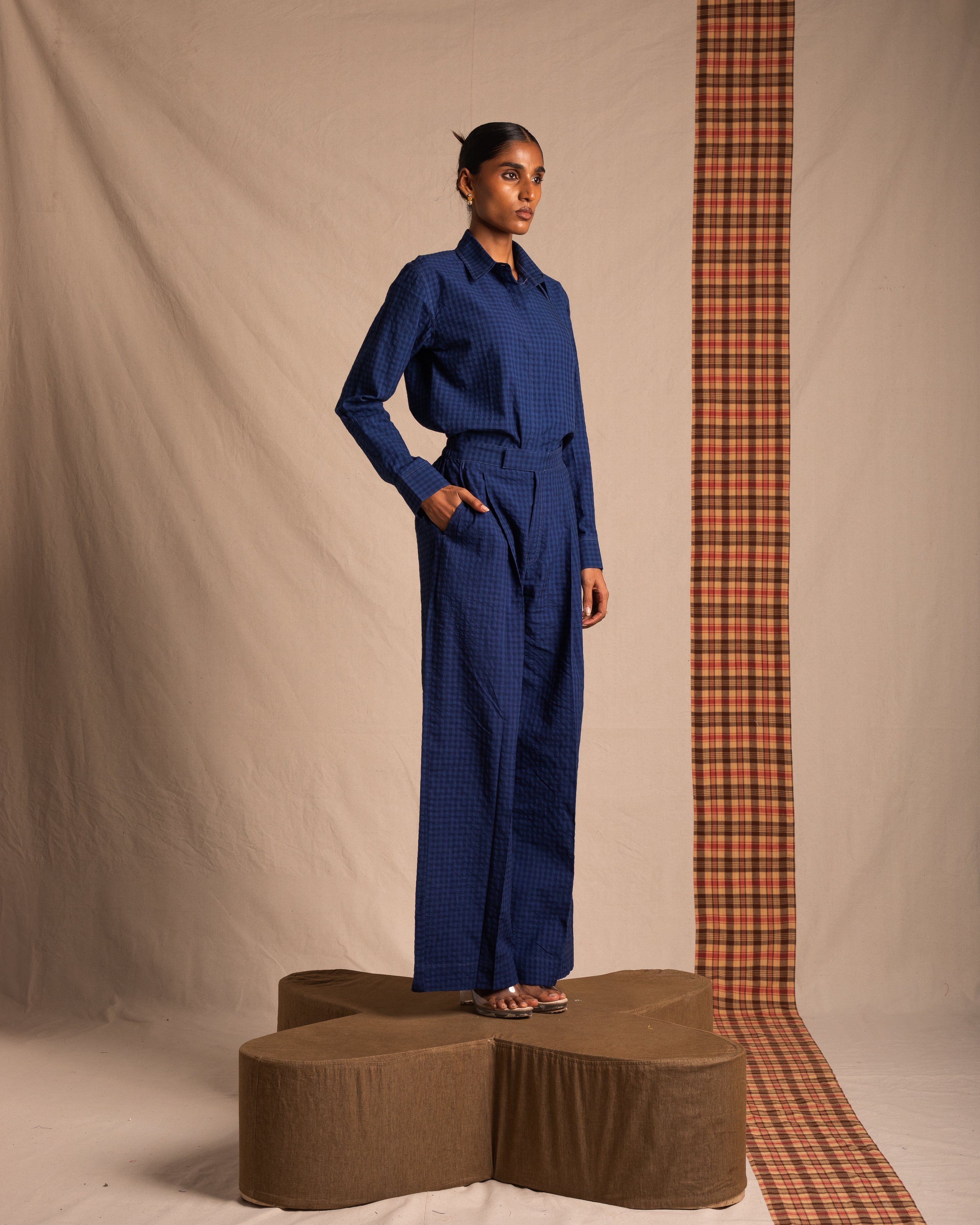 Woman in blue checkered jumpsuit standing on brown platform with plaid backdrop, fashion photoshoot