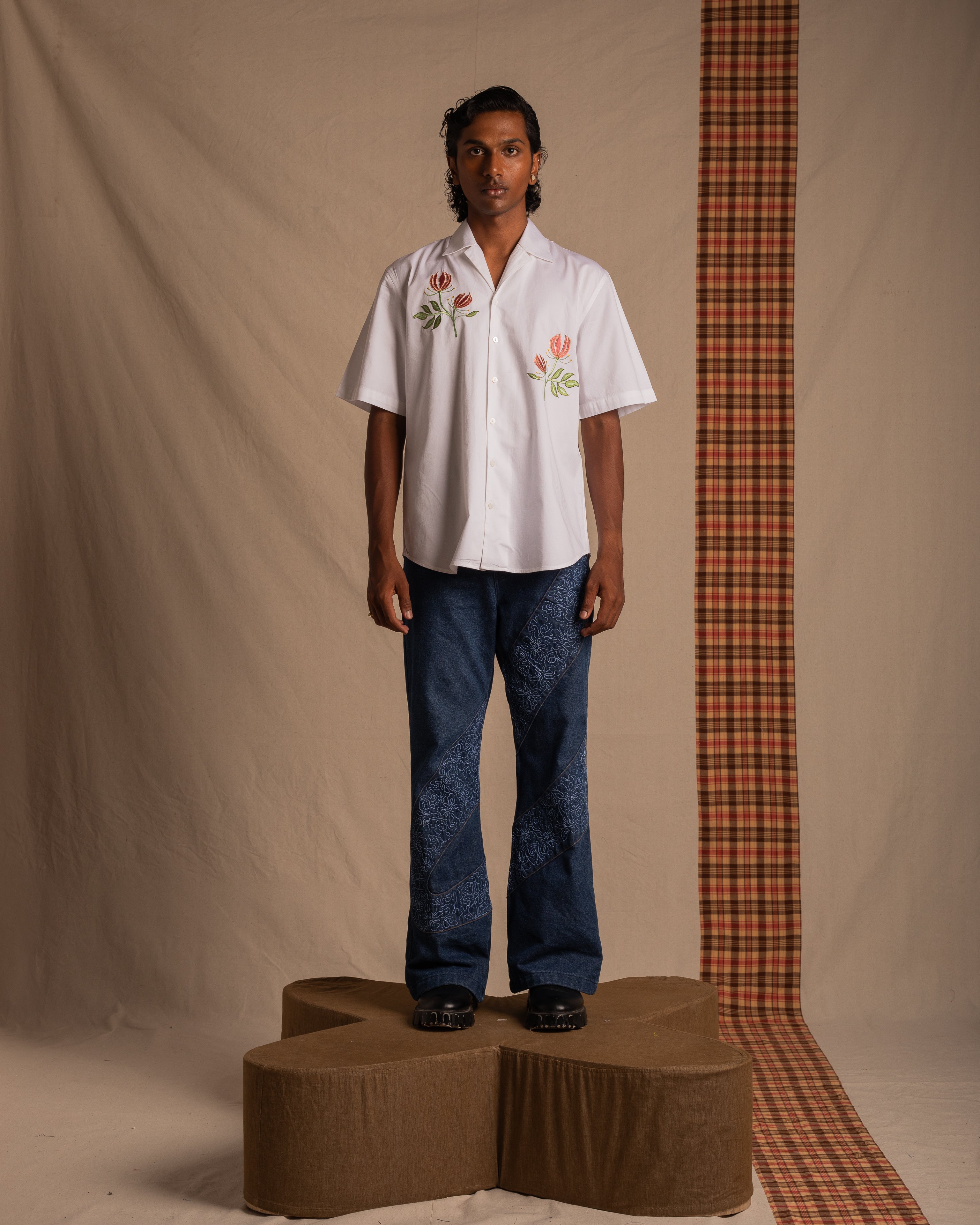 Man in floral embroidered white short-sleeve shirt and patterned blue jeans, standing on a platform in a streetwear fashion photoshoot