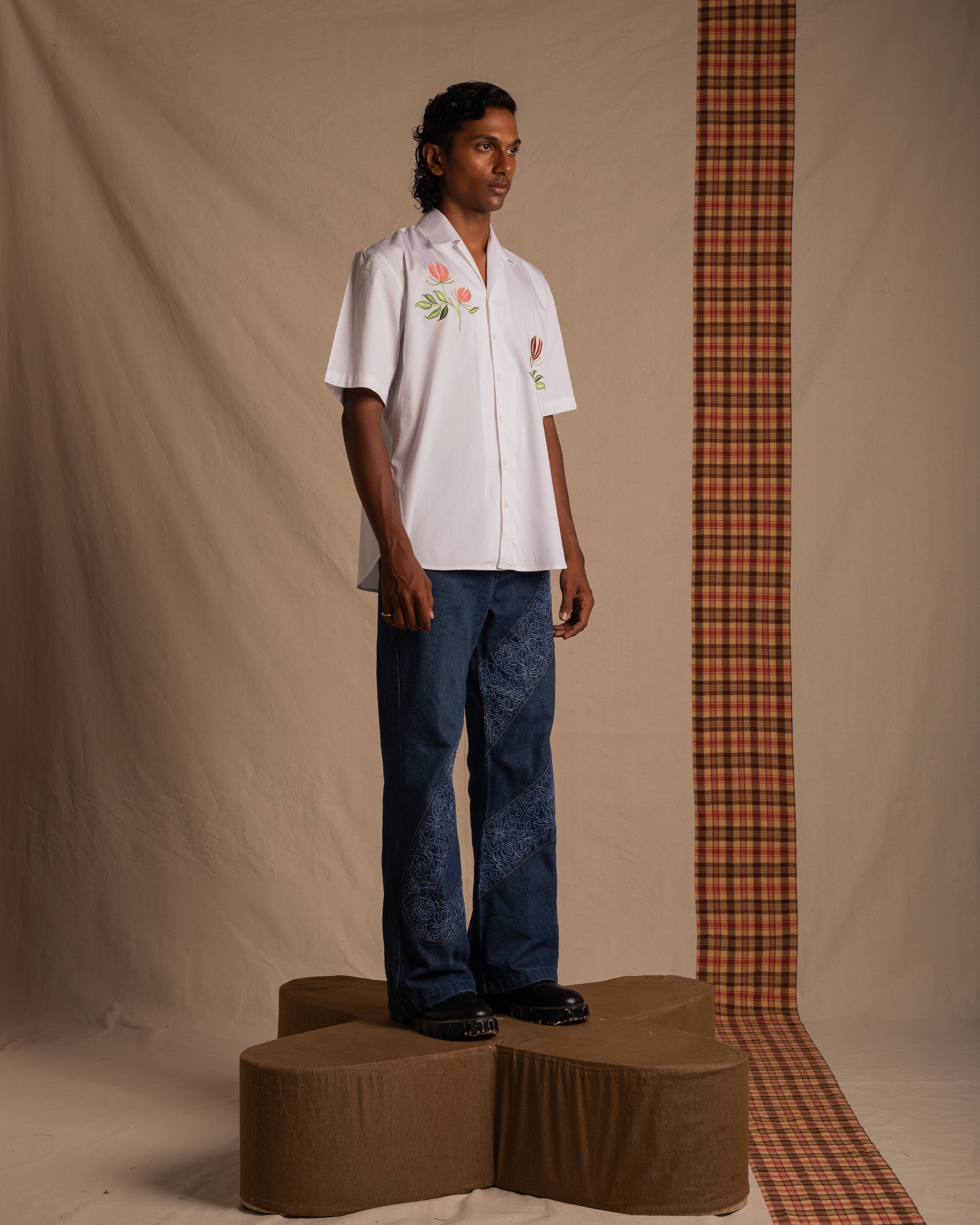 Male model wearing a white floral short sleeve shirt and blue patterned jeans, standing on a brown platform in a studio with a beige and plaid backdrop, streetwear fashion style.