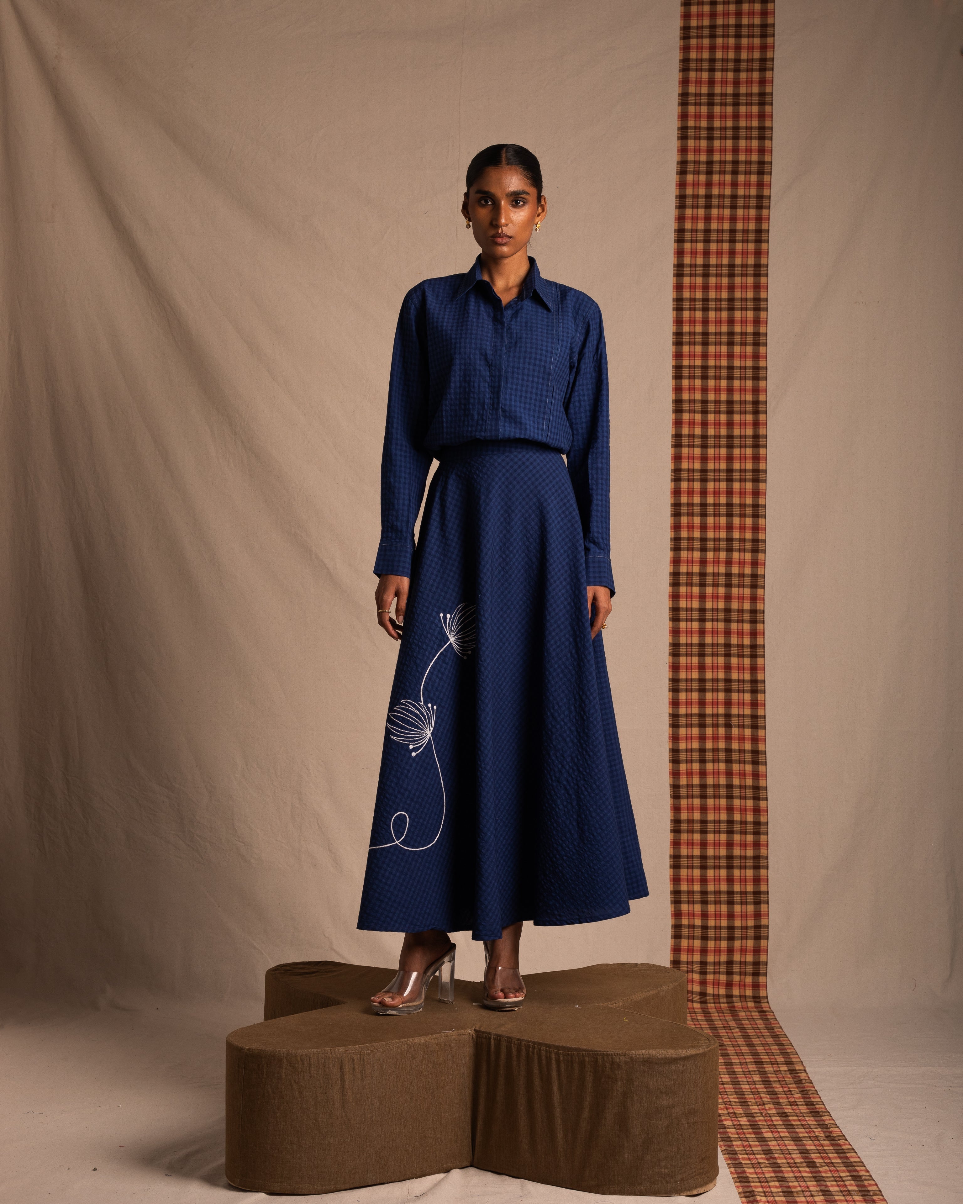 Woman in a blue textured shirt and matching long skirt with white embroidery, standing indoors.