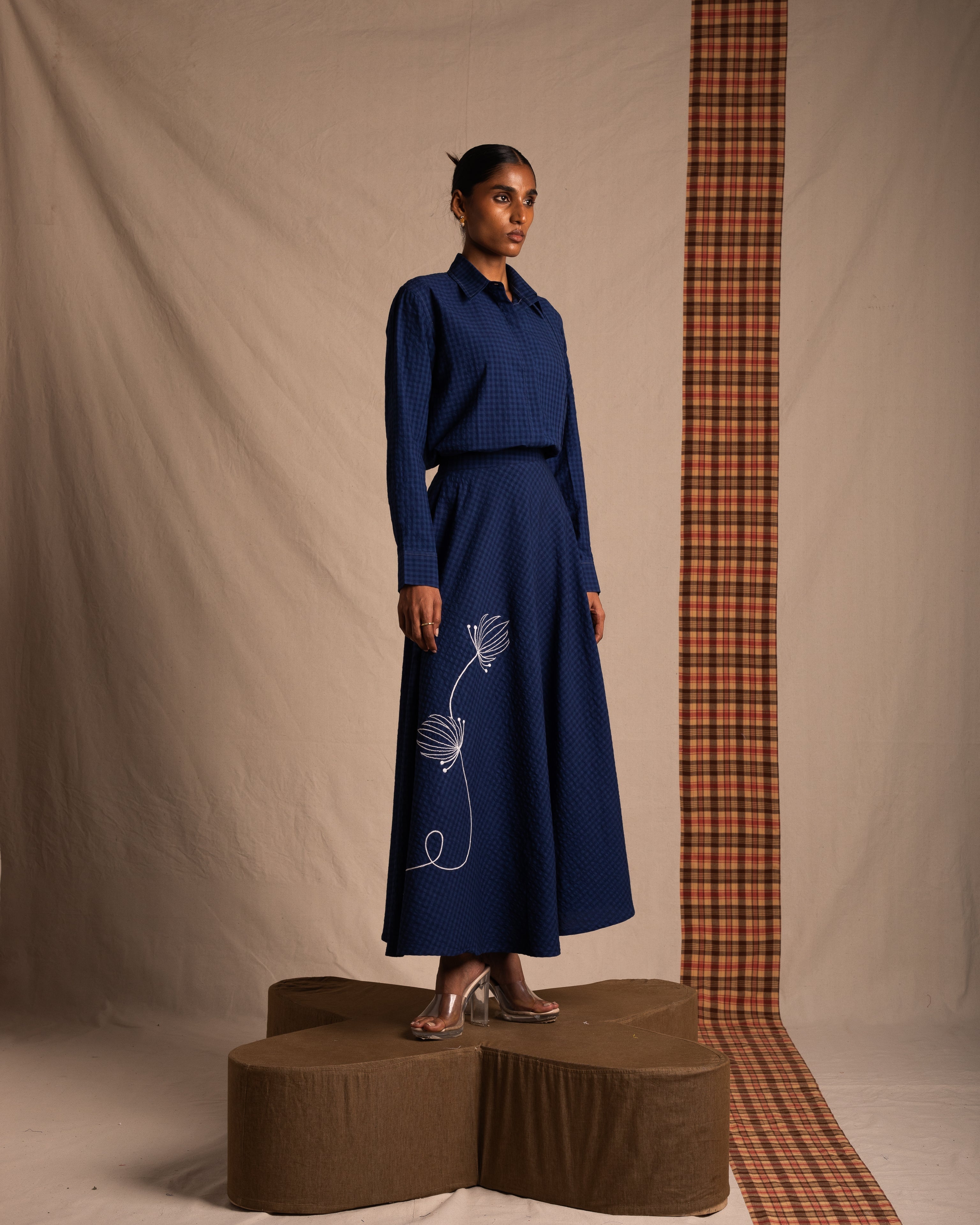 Woman modeling a blue textured long-sleeve blouse and matching maxi skirt with white floral design accents, standing on a platform in a minimalist studio setting, stylish streetwear fashion.