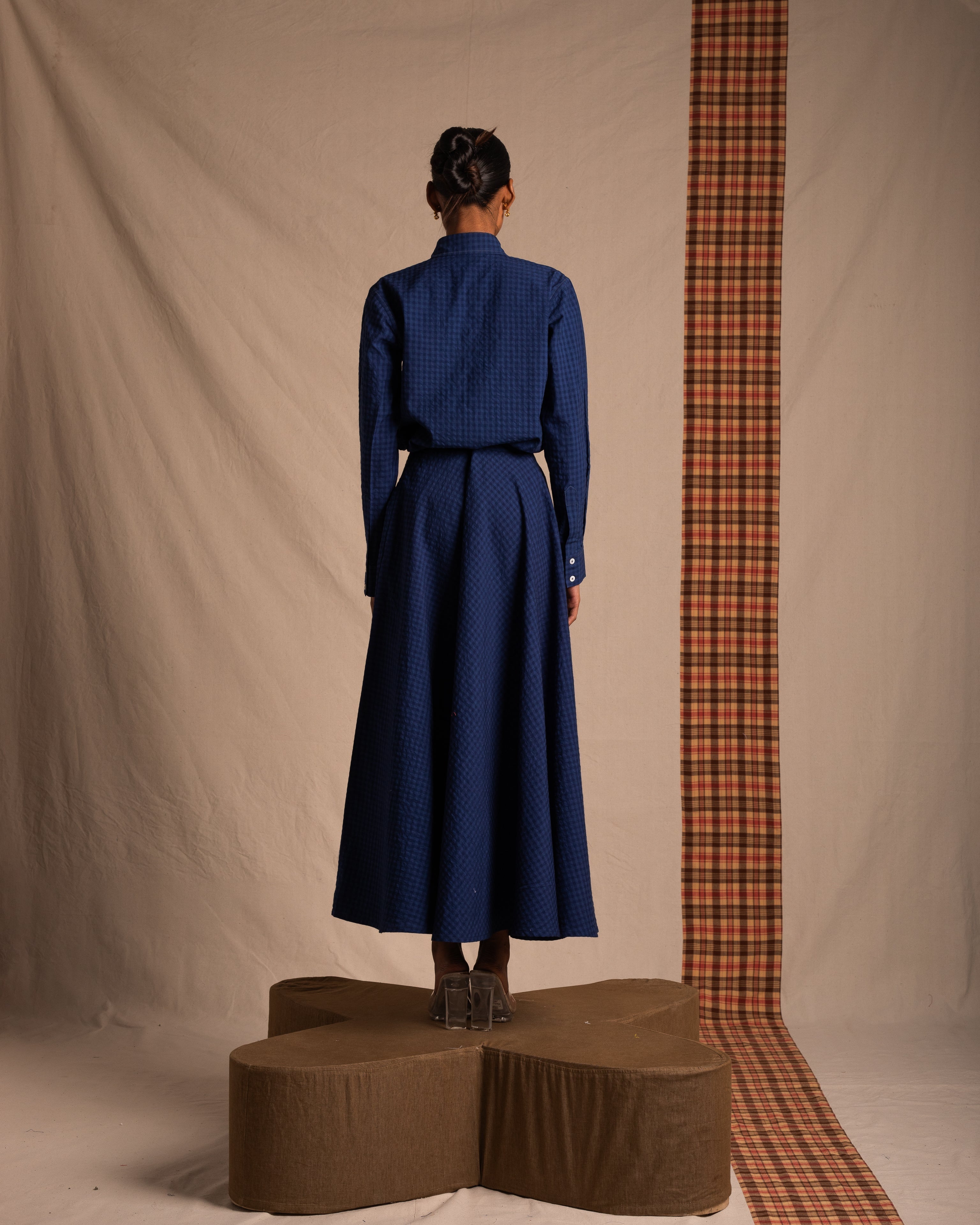 Woman in blue textured long dress, standing on brown platform with plaid accent backdrop.