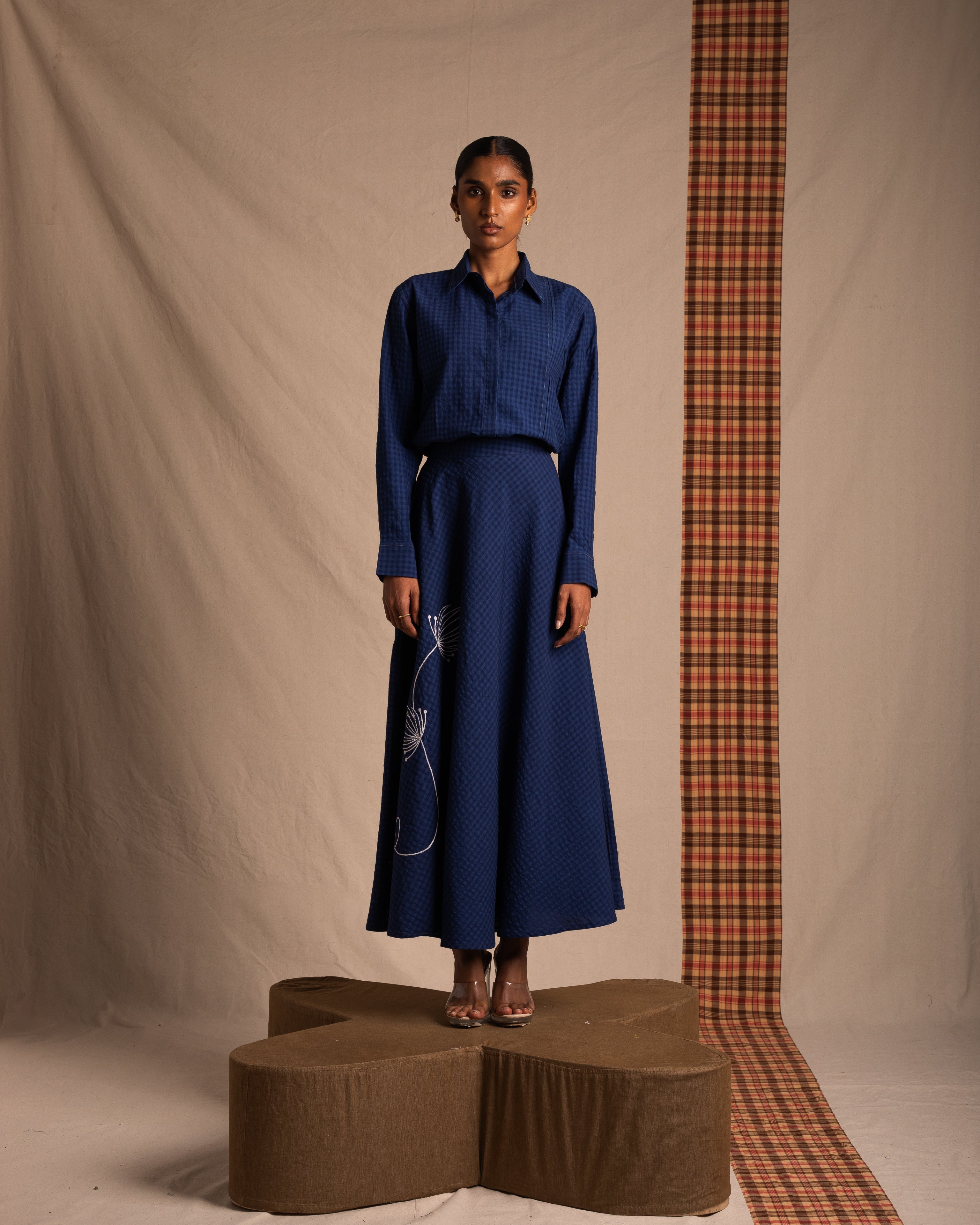 Woman modeling a long blue textured skirt and matching collared shirt, standing indoors.