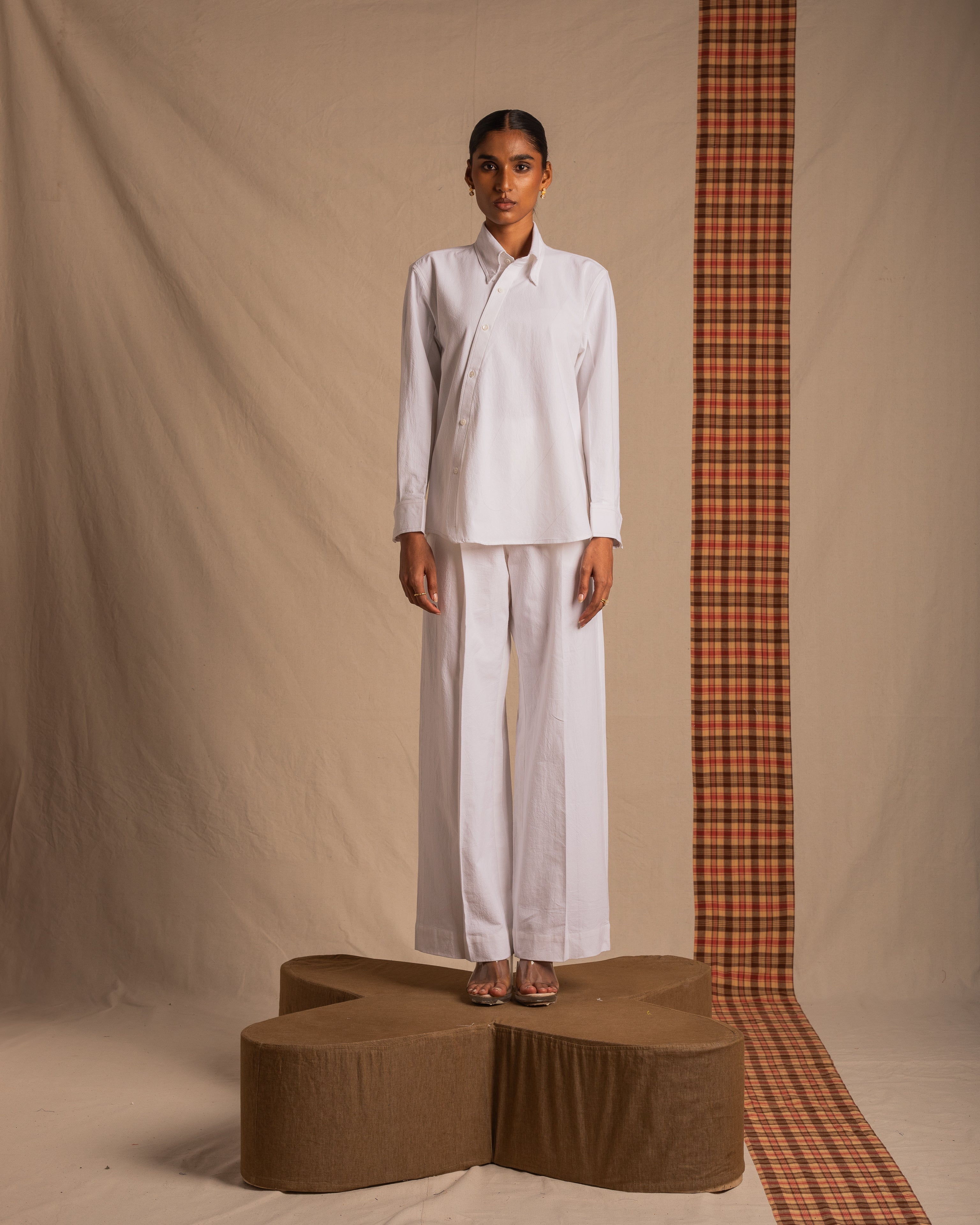 Woman modeling white button-up shirt and wide-leg pants, standing on a brown platform against a beige backdrop.