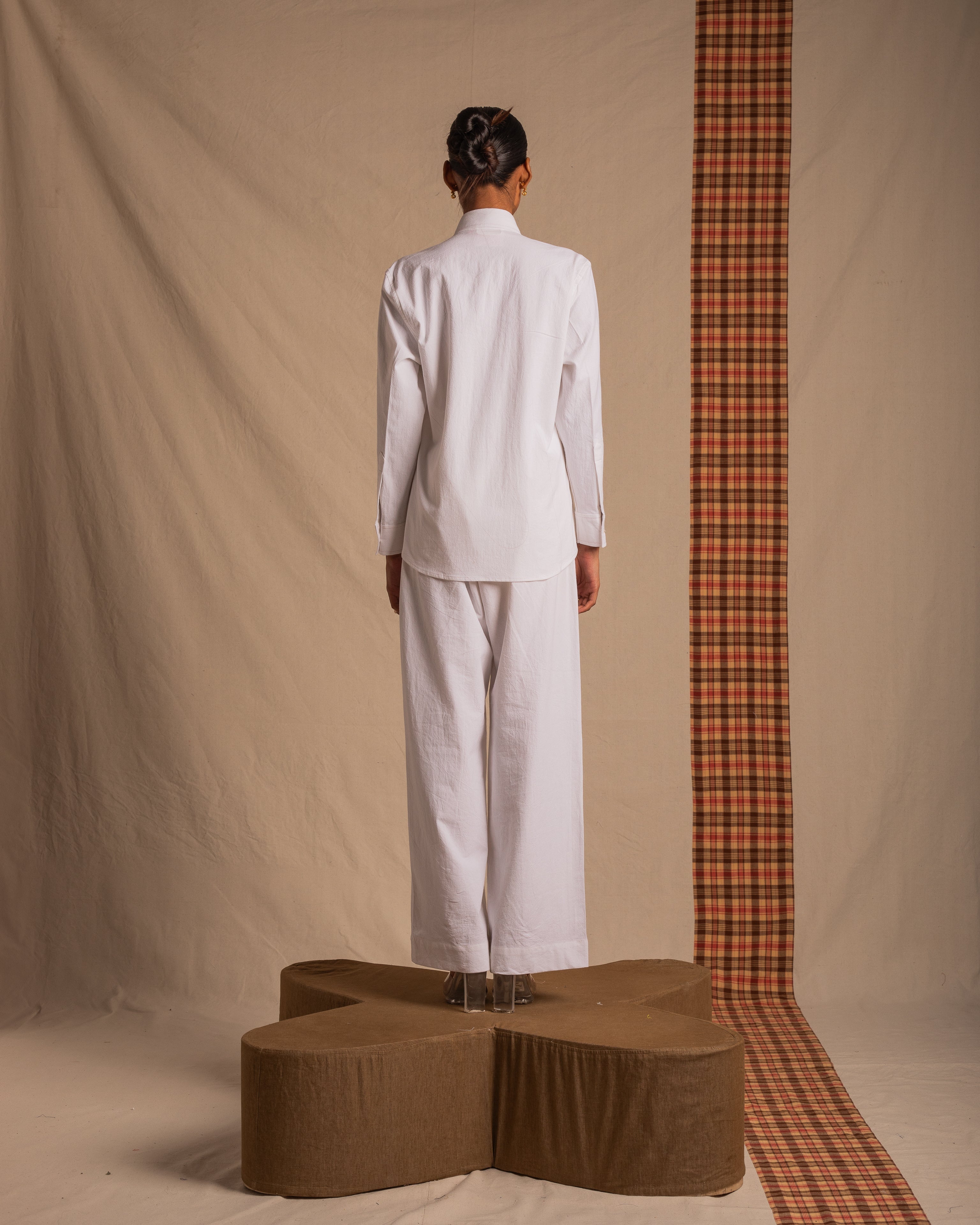 Model in white long sleeve shirt and wide-leg pants stands on a platform, studio background.