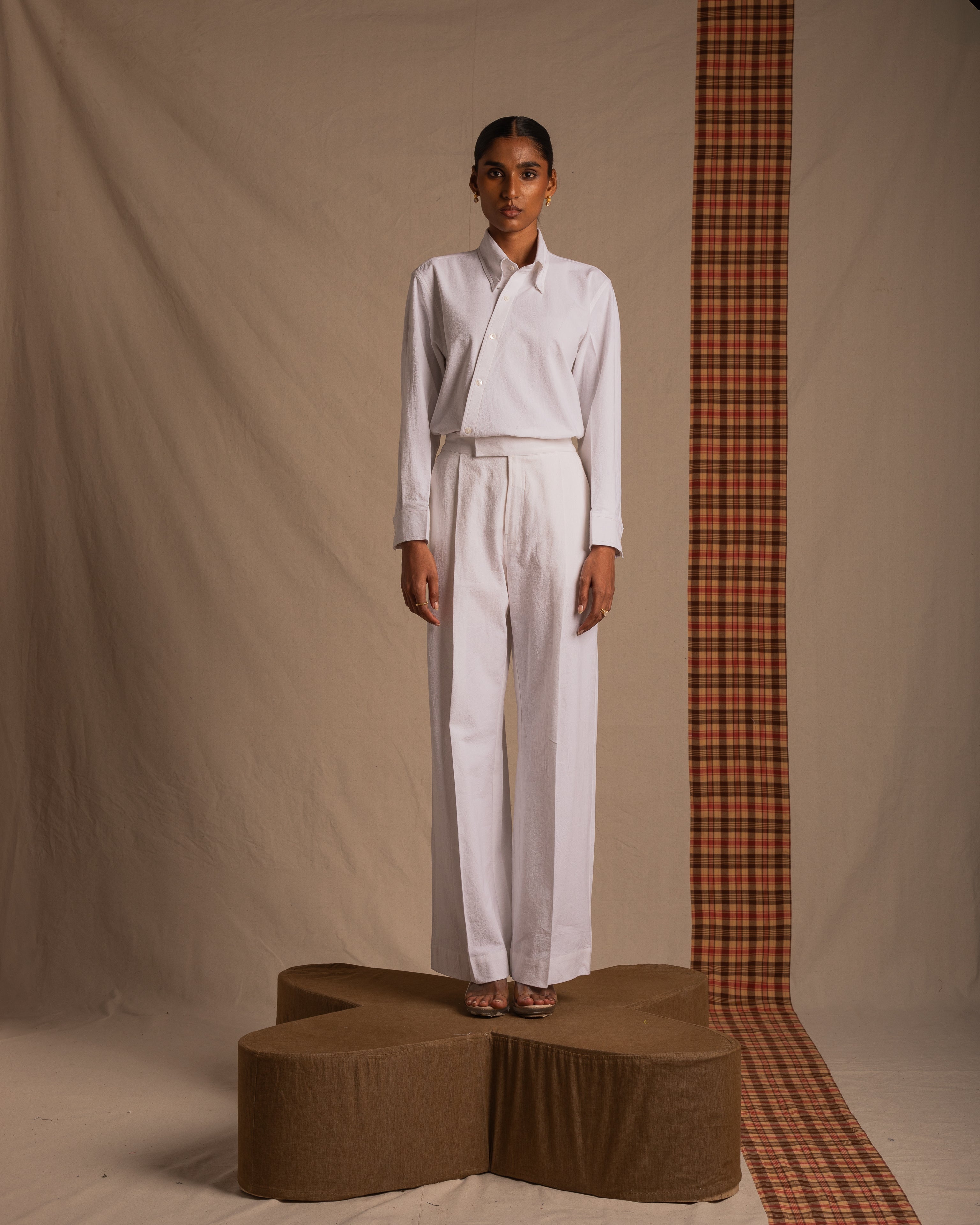Woman in white button-up shirt and wide-leg trousers stands on brown platform, neutral background