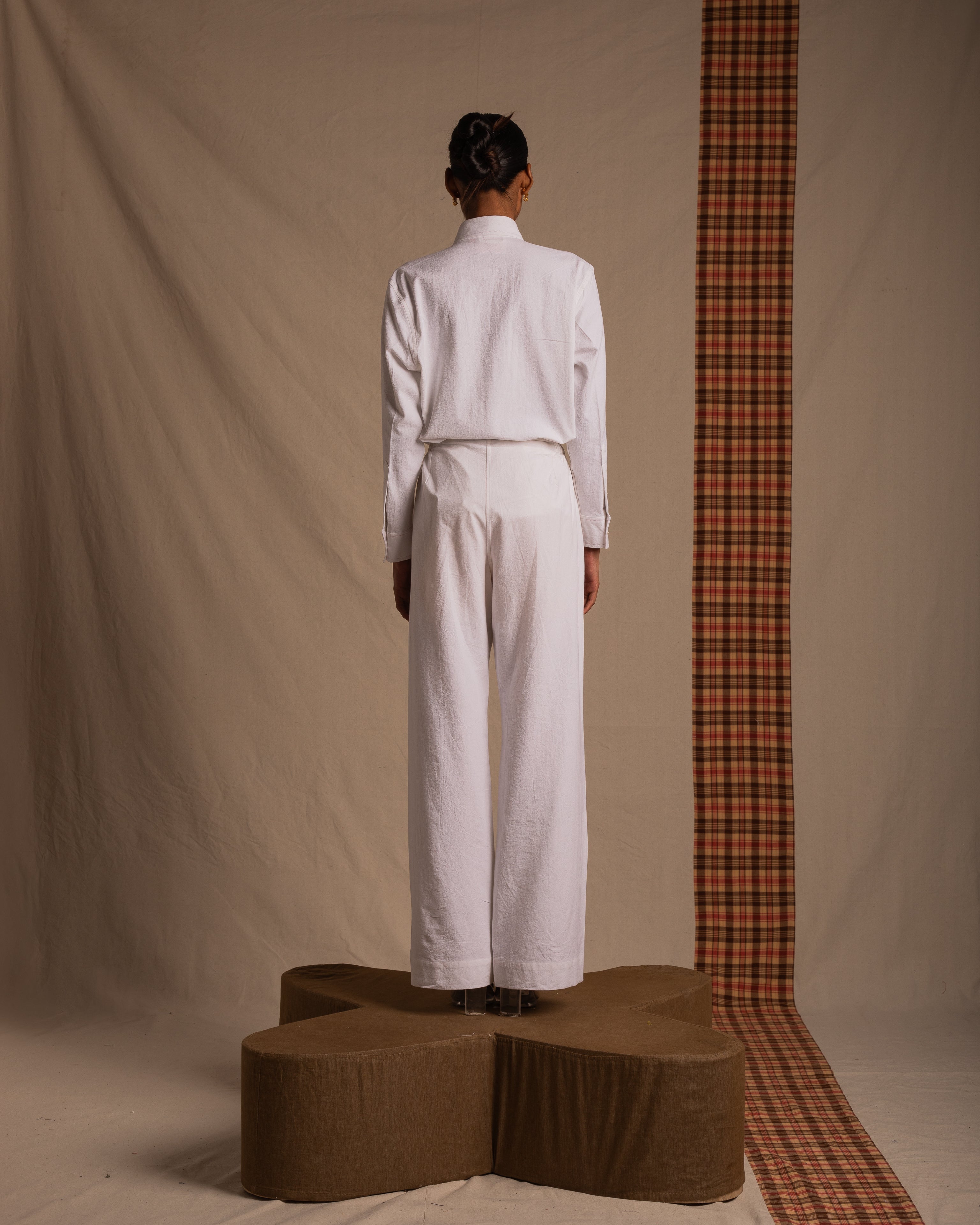 Model in white shirt and wide-leg pants stands on brown platform with plaid fabric backdrop