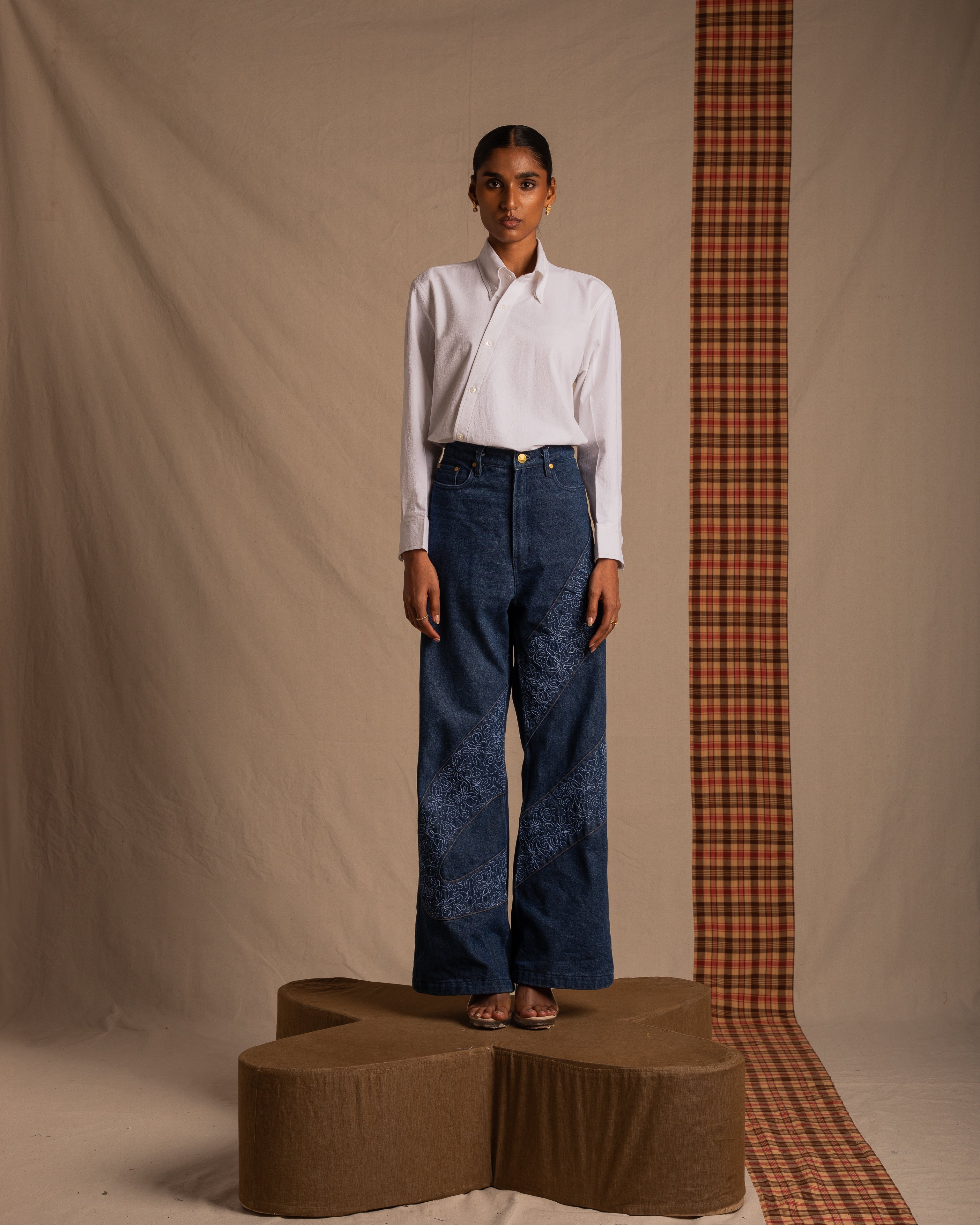 Woman in white shirt and wide-leg embroidered jeans stands on brown platform, minimalist studio background.