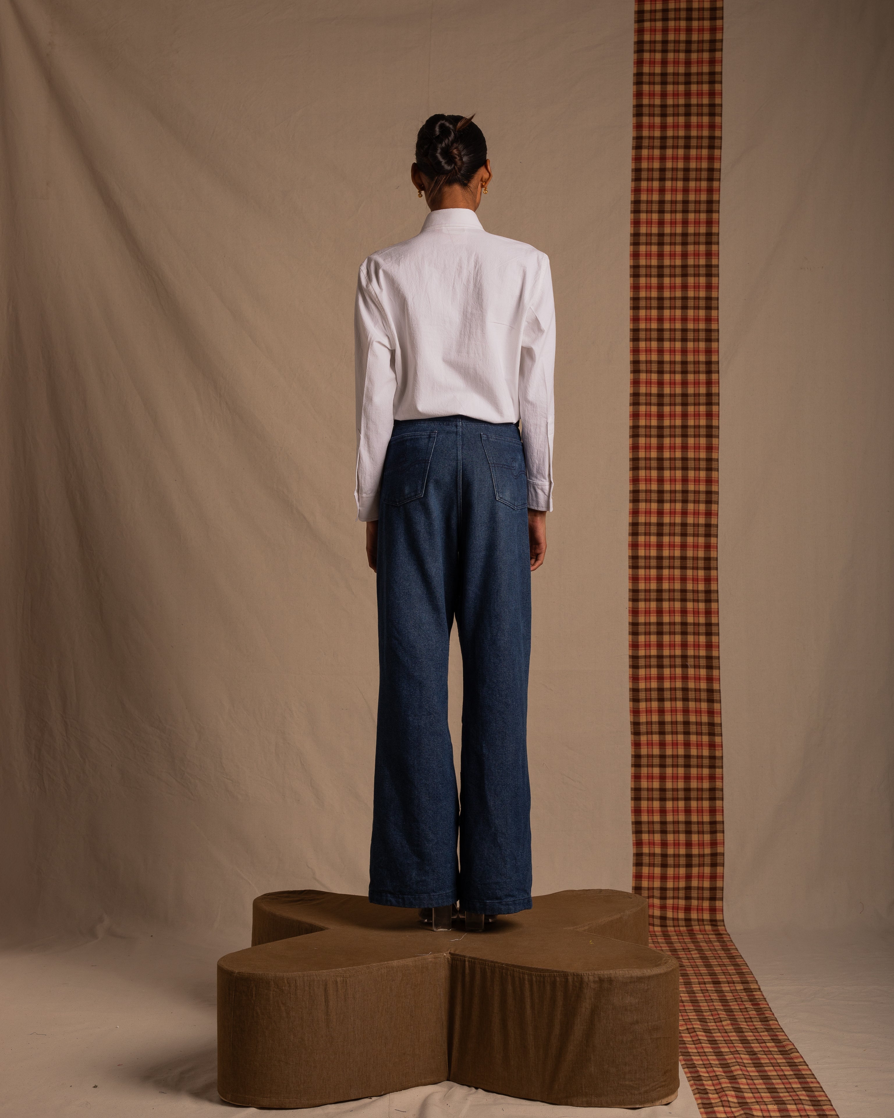 Model in white shirt and wide-leg blue jeans, standing on a platform, neutral studio backdrop.