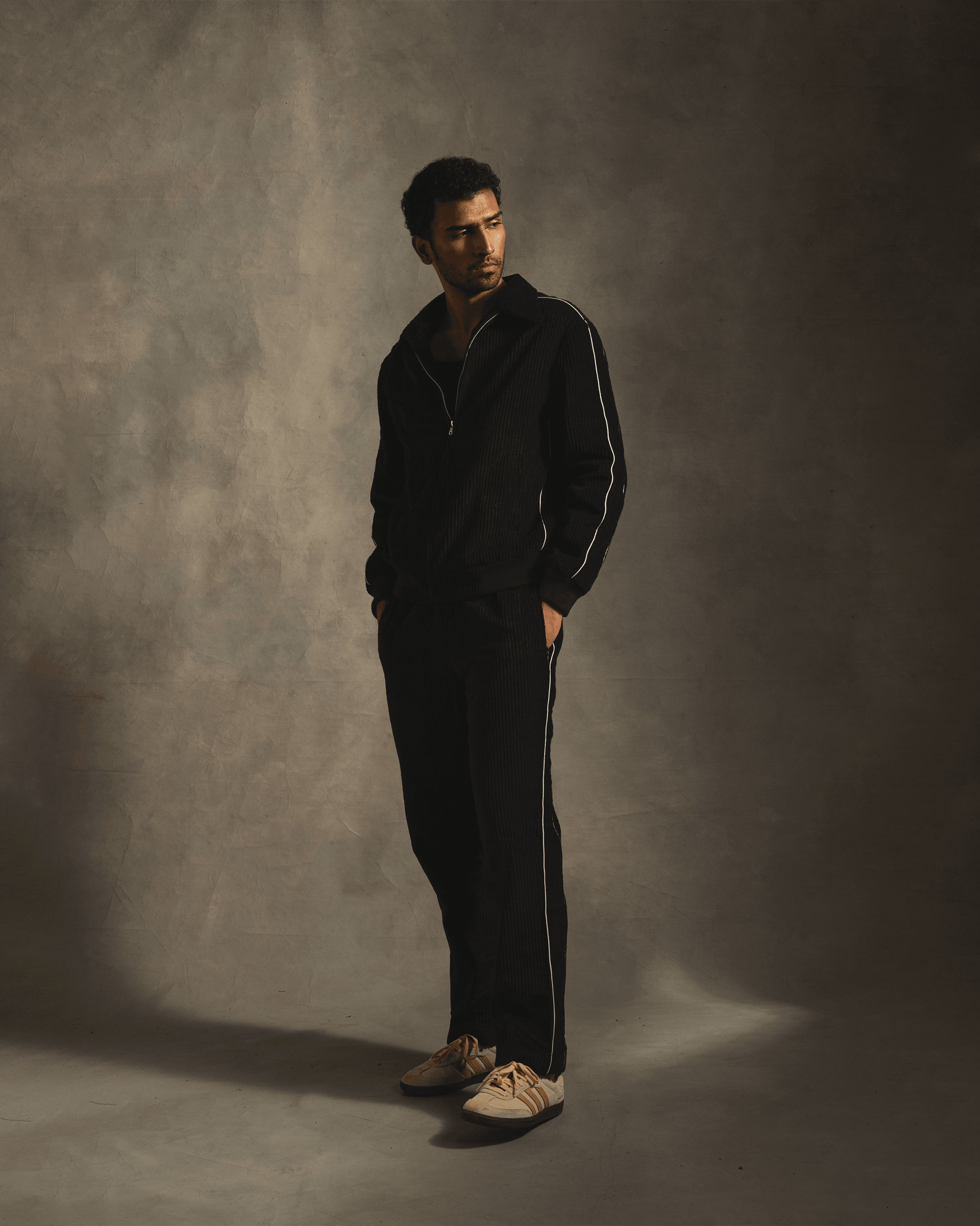 Man wearing black streetwear tracksuit with white stripes and beige sneakers, neutral background