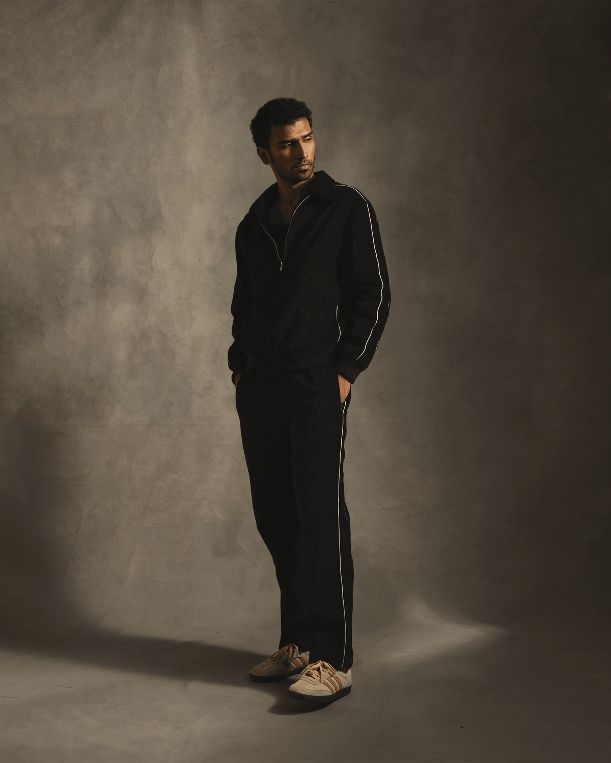 Man wearing black streetwear tracksuit with white stripes and beige sneakers, neutral background