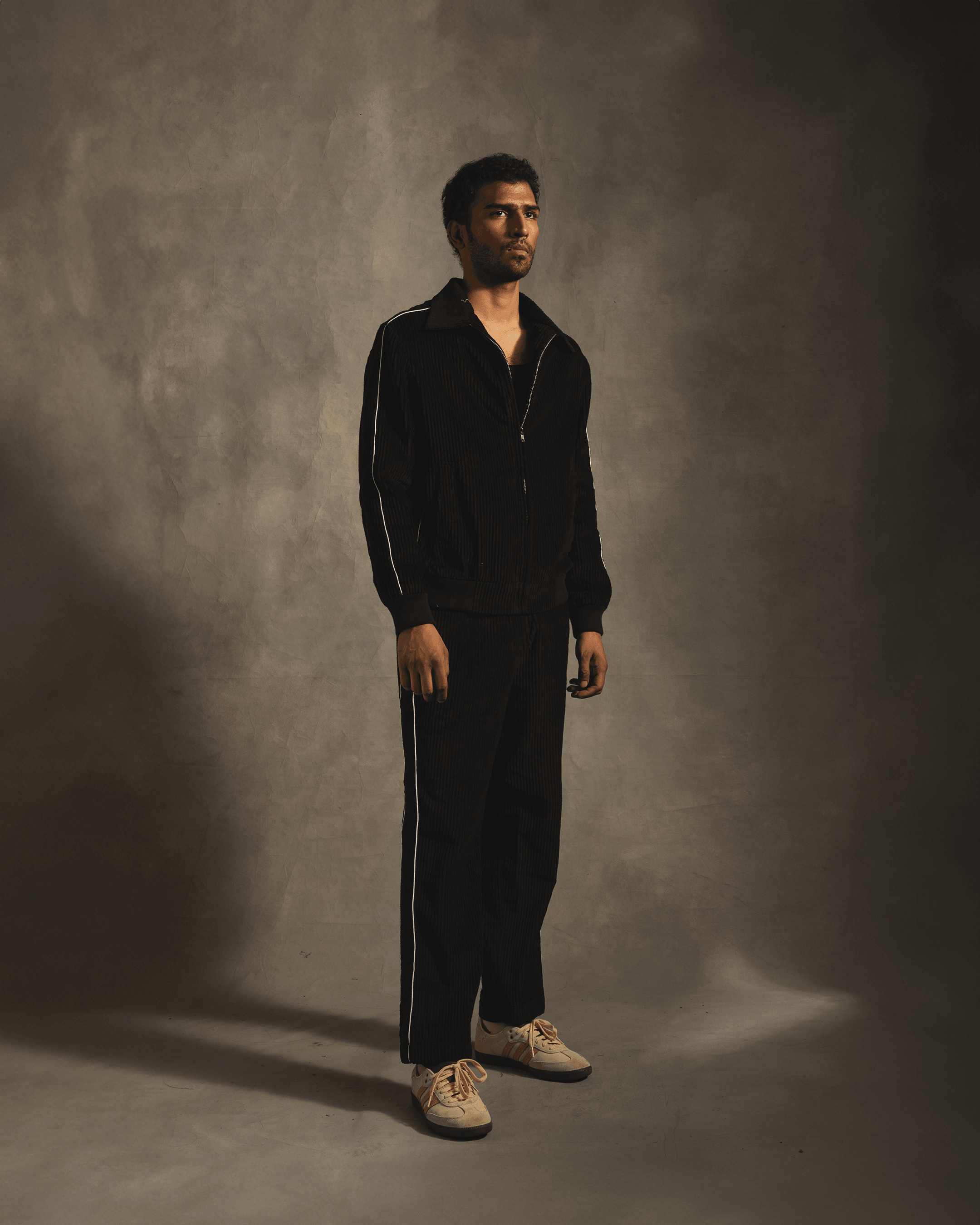 Man wearing black tracksuit with white side stripes and white sneakers, studio background