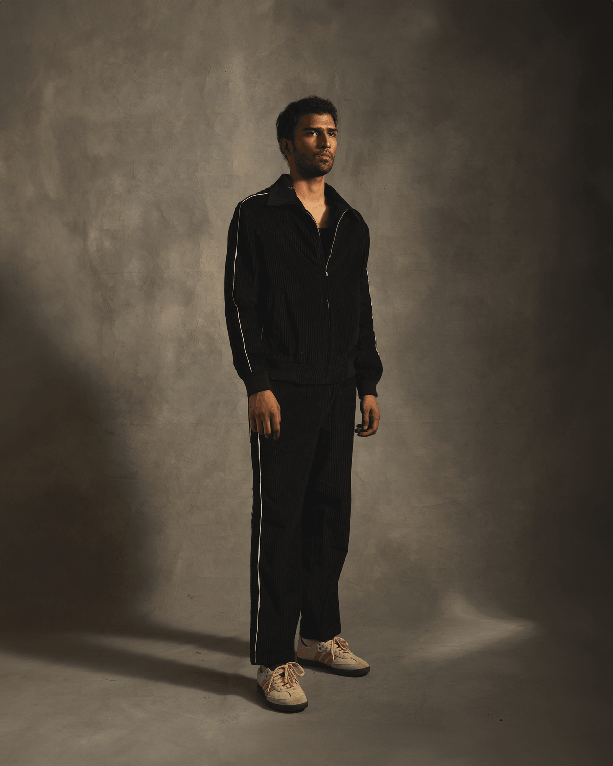 Man wearing black tracksuit with white side stripes and white sneakers, studio background