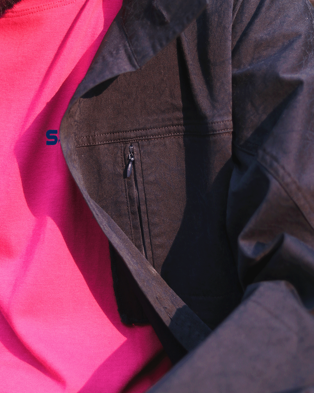 Close-up of black streetwear jacket with zipper pocket over bright pink t-shirt outdoors.