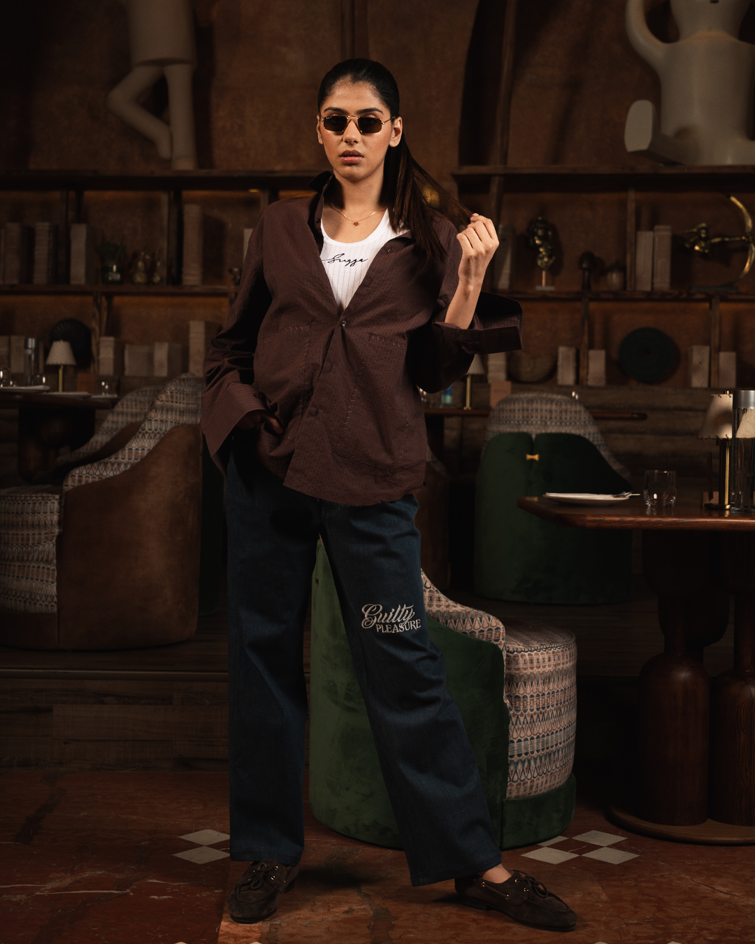 Woman in brown shirt, white graphic tee, and blue embroidered jeans posing indoors in modern lounge.