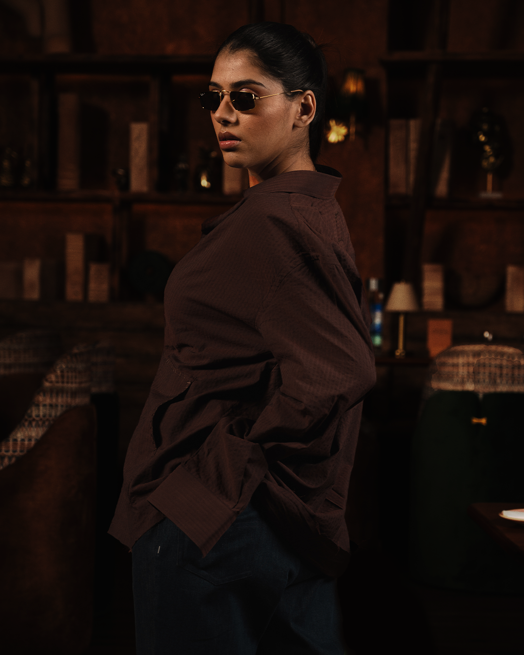 Woman in brown oversized shirt and sunglasses posing indoors, streetwear fashion style