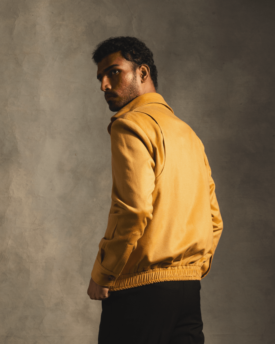 Man wearing a tan cashmere bomber jacket with black pants against a gray textured background