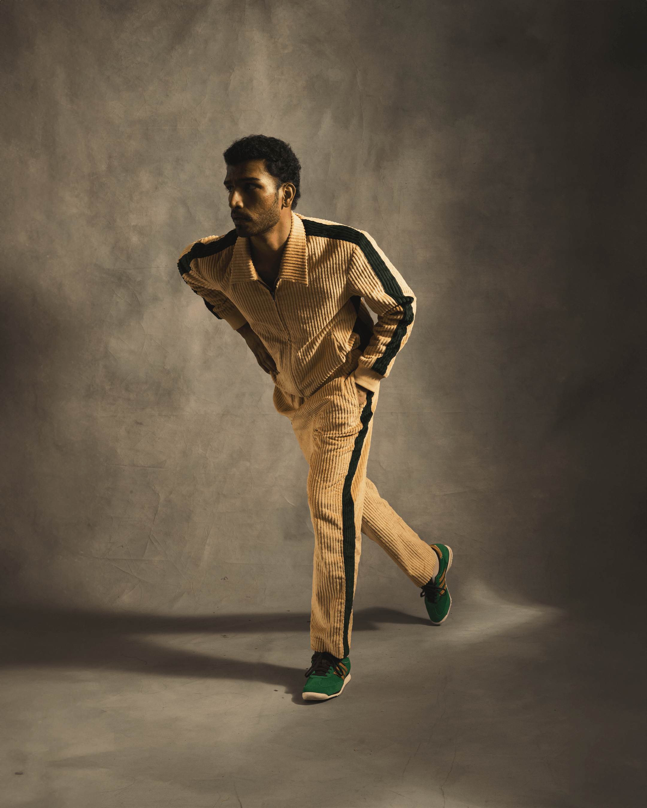Man modeling a cream corduroy tracksuit with green stripes and green sneakers, studio lit