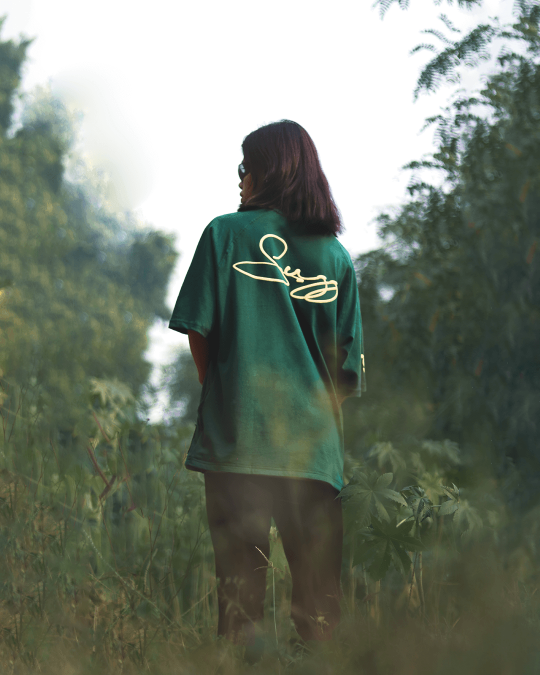 Person in green streetwear T-shirt with signature print standing outdoors in greenery