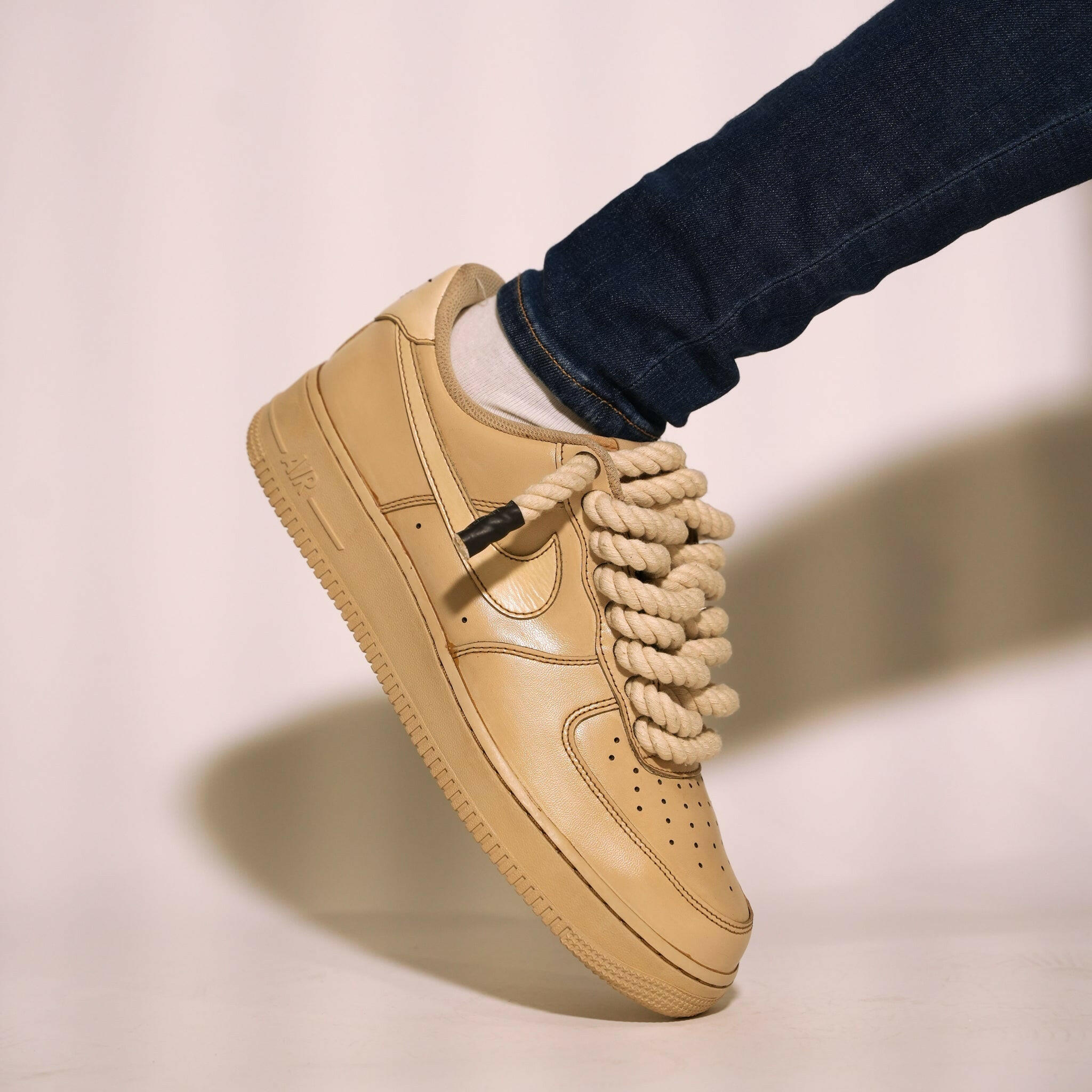 Tan streetwear sneaker with oversized rope laces, worn with dark jeans and white socks