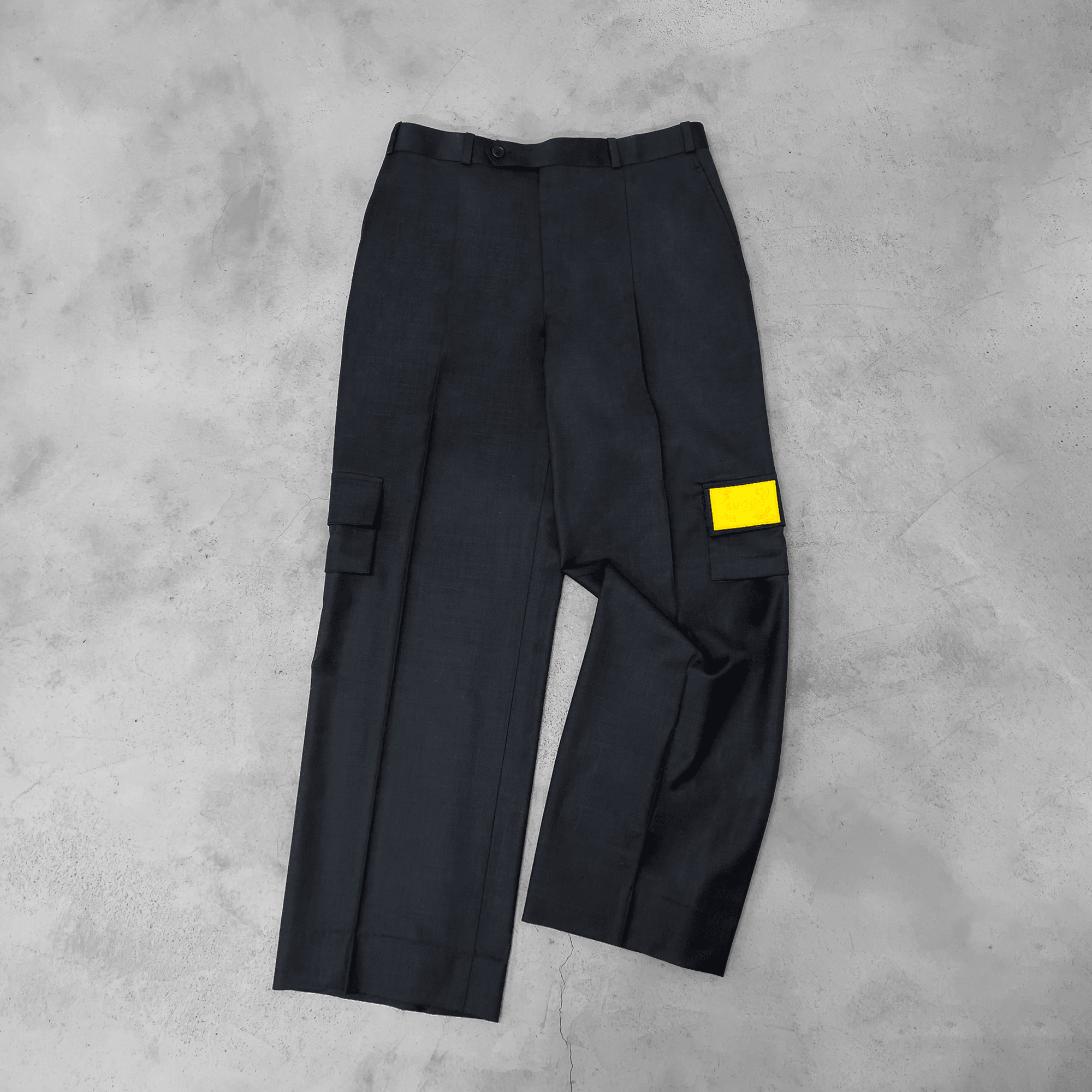 Dark grey cargo pants with yellow patch detail on side pocket, laid flat on a concrete floor