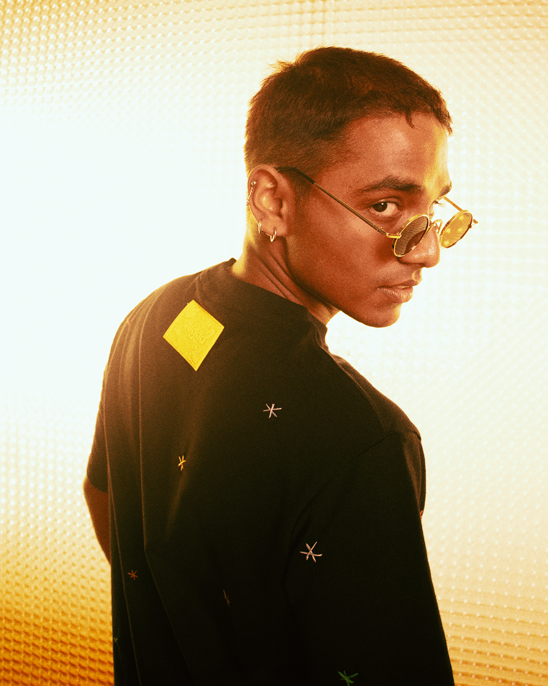 Streetwear model in black graphic tee with yellow patch and sunglasses under warm lighting