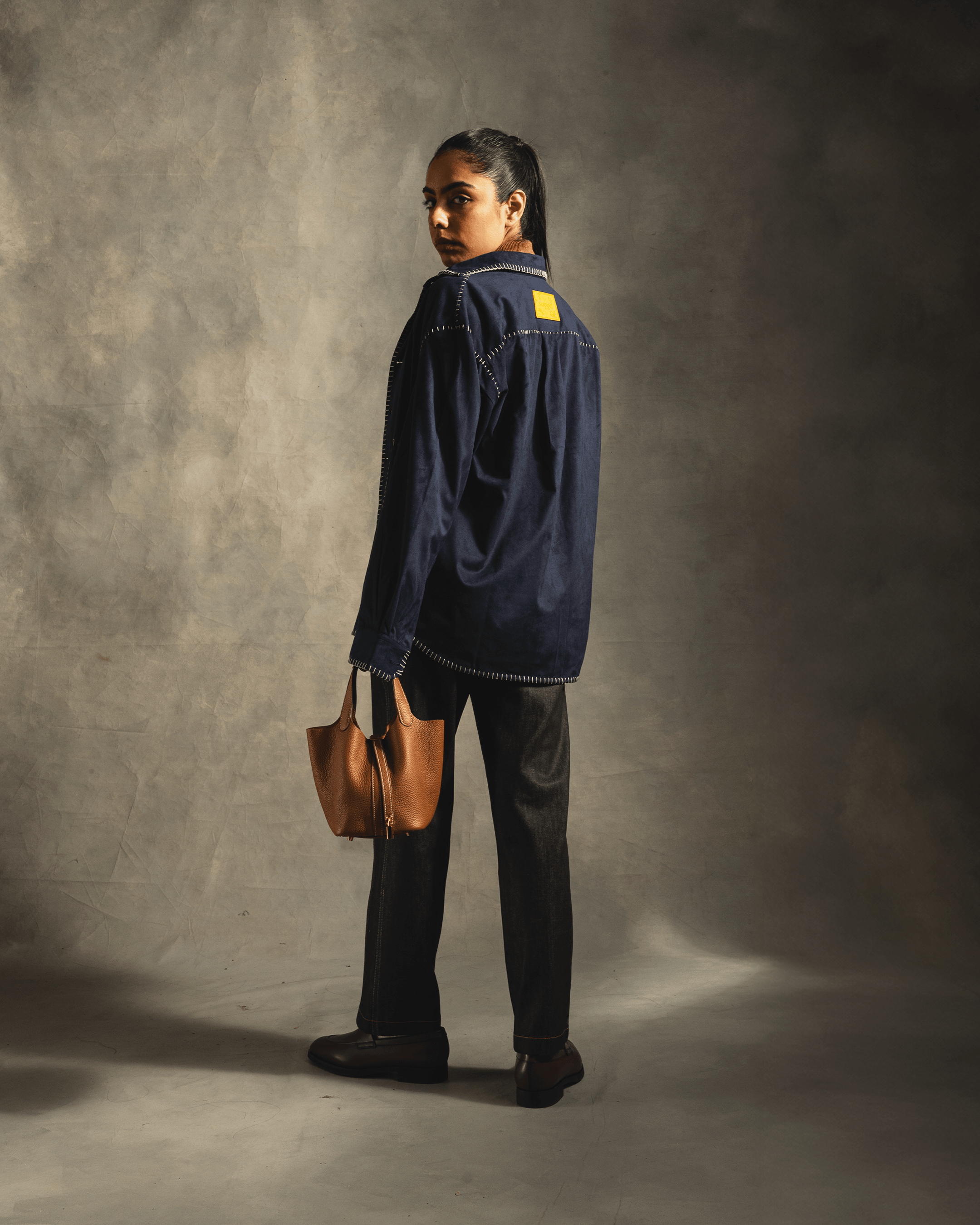 Woman in navy streetwear jacket with visible flatlock stitching and yellow patch, holding a tan leather tote bag, facing away in a studio setting.