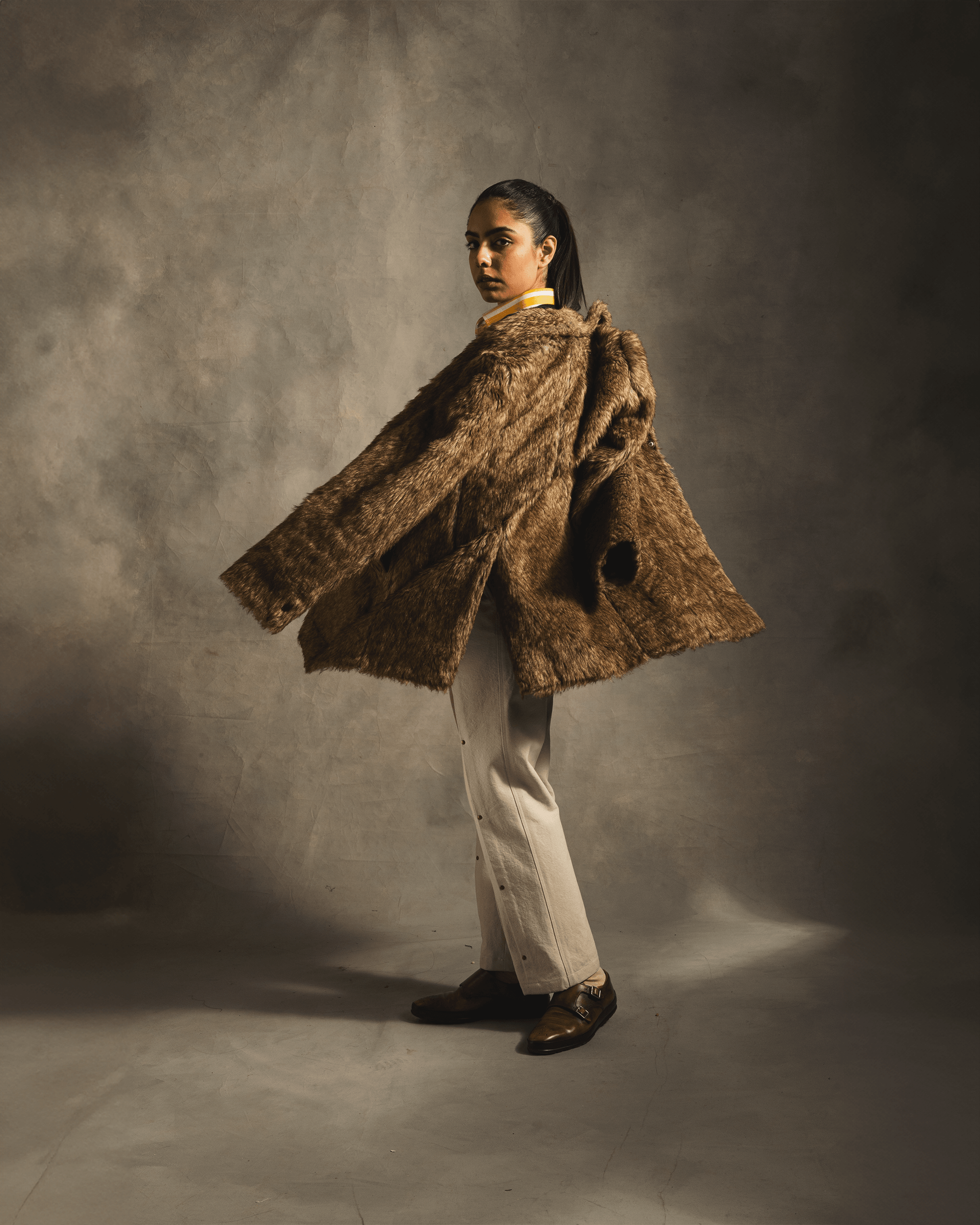 Model wearing oversized brown faux fur coat, cream pants, and loafers in studio fashion shot