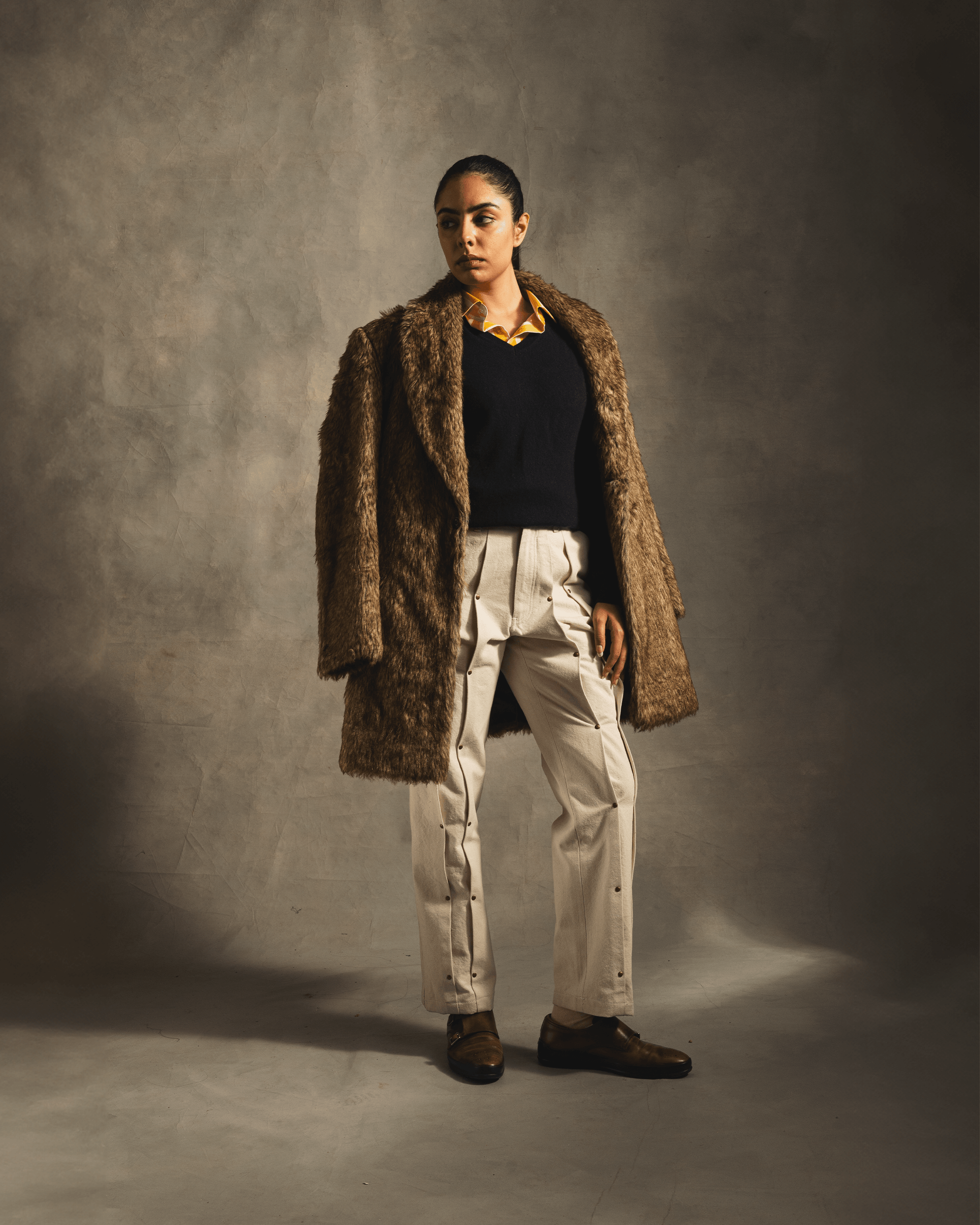 Woman in brown faux fur coat, black sweater, cream pants, and brown shoes, studio shot