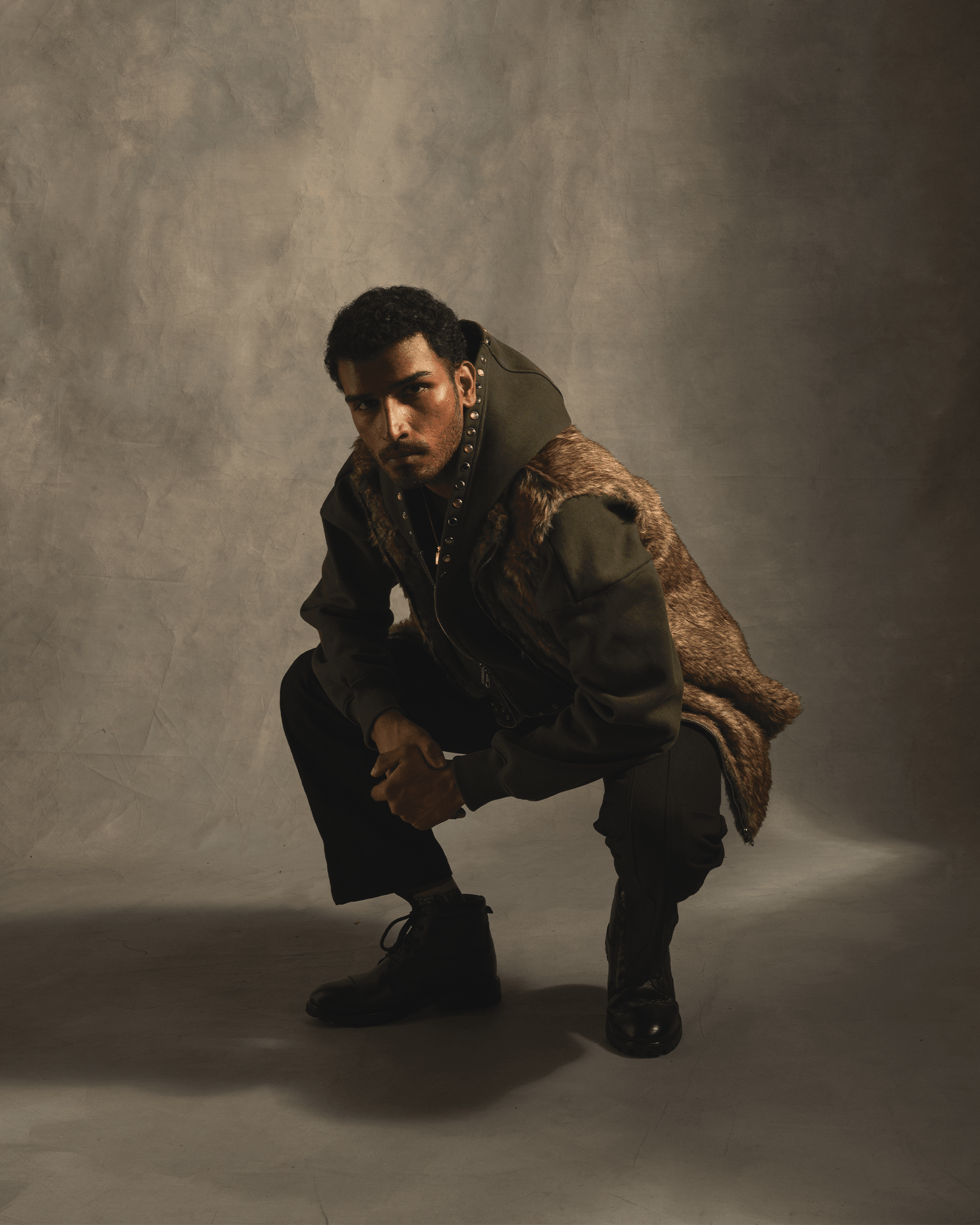 Man in fur-trimmed streetwear jacket and black boots squatting against neutral background