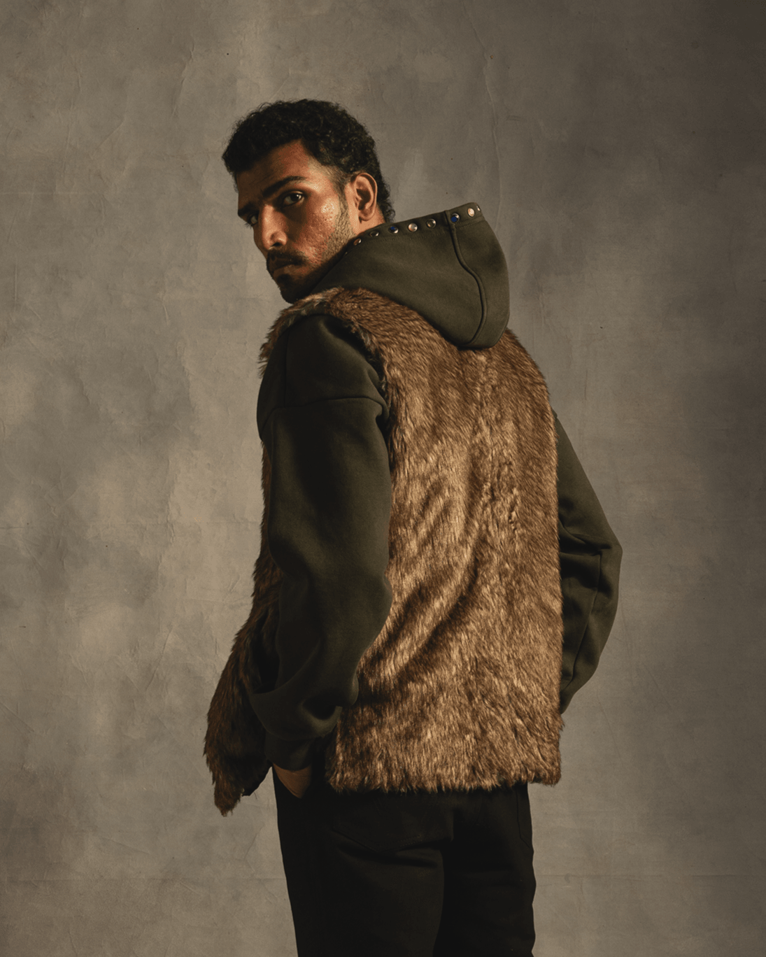 Man in a faux fur vest over a dark hoodie, streetwear fashion, neutral backdrop