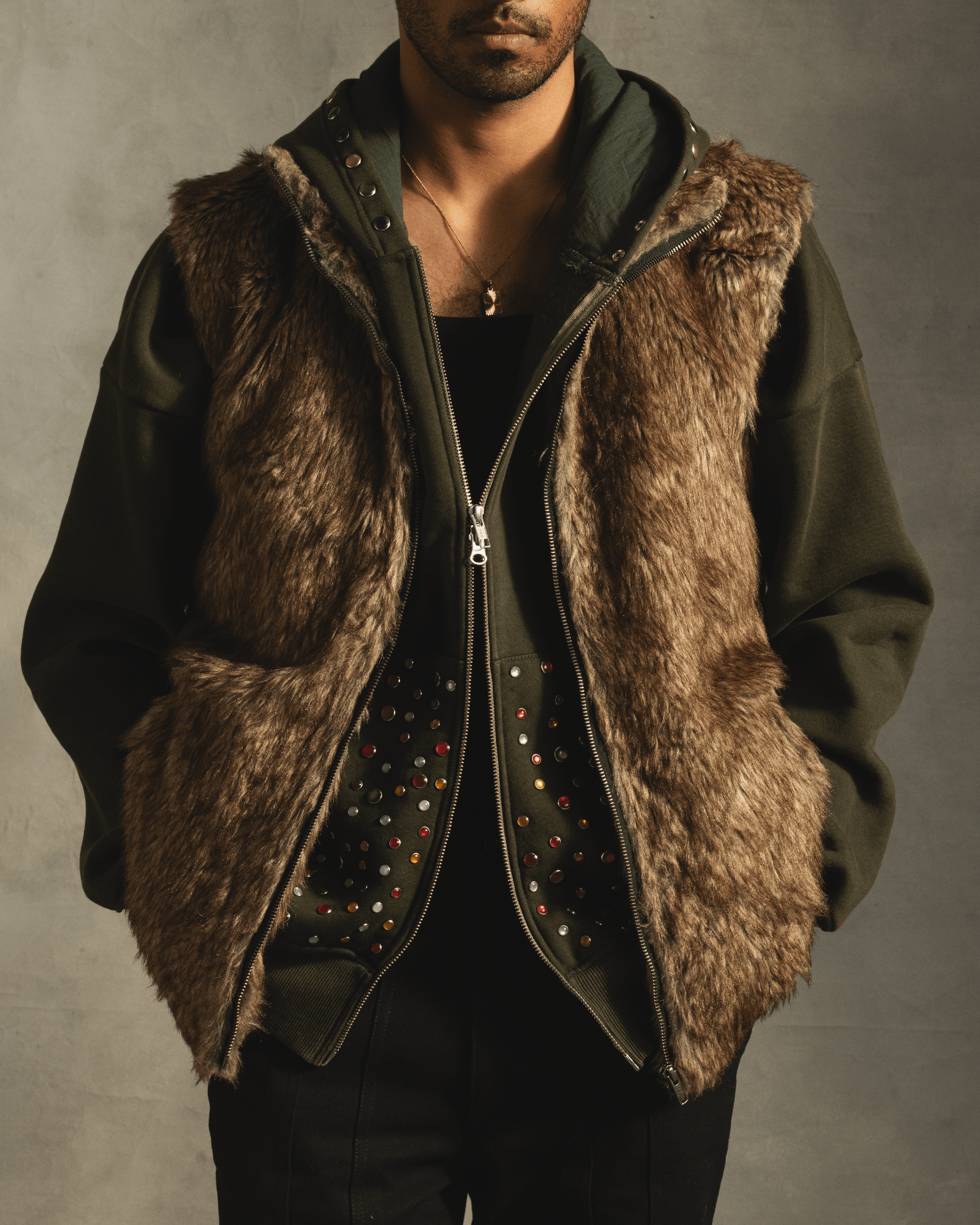 Man wearing faux fur vest layered over studded dark green hoodie, streetwear fashion