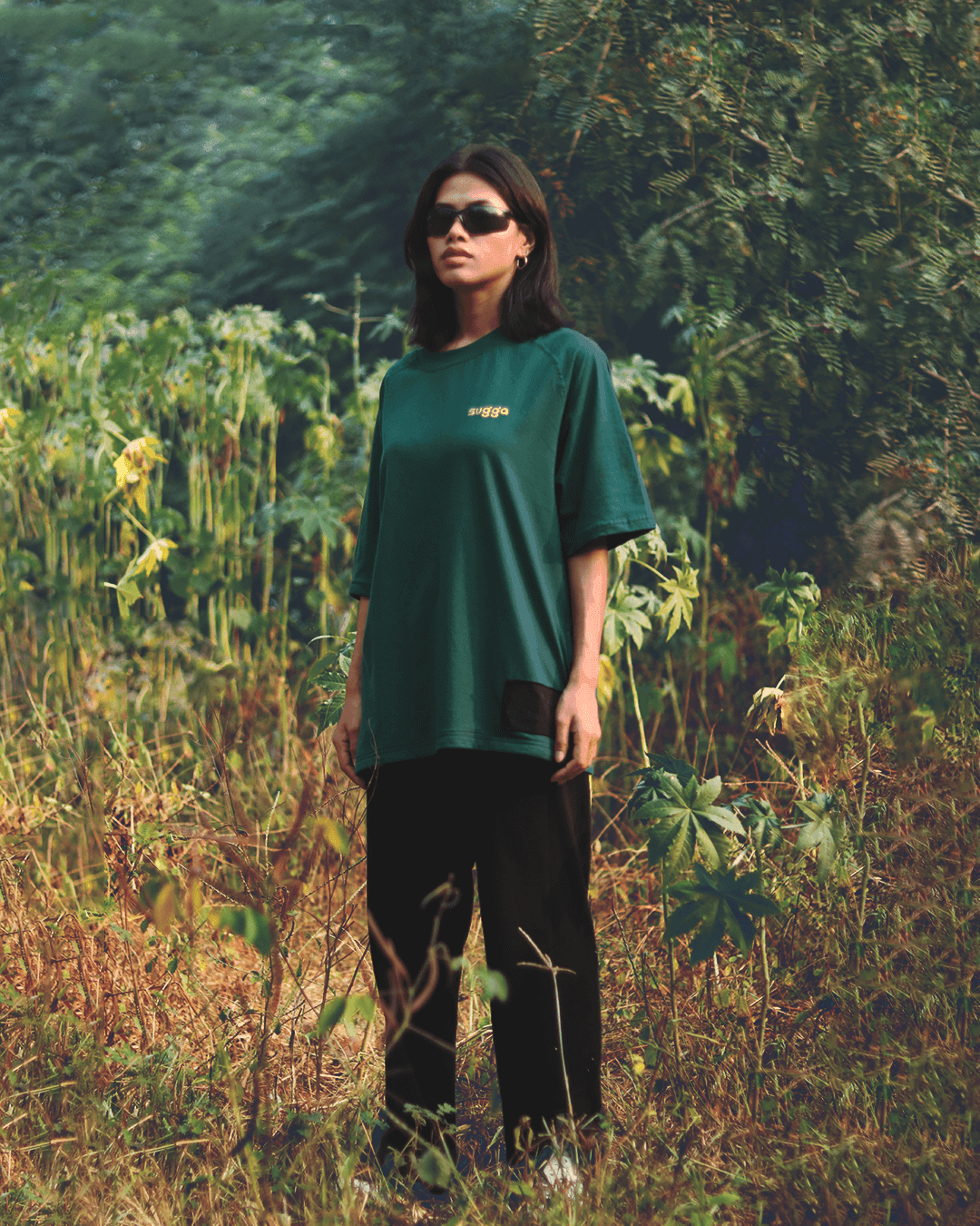Woman wearing a dark green streetwear t-shirt and black pants, posing outdoors in greenery.