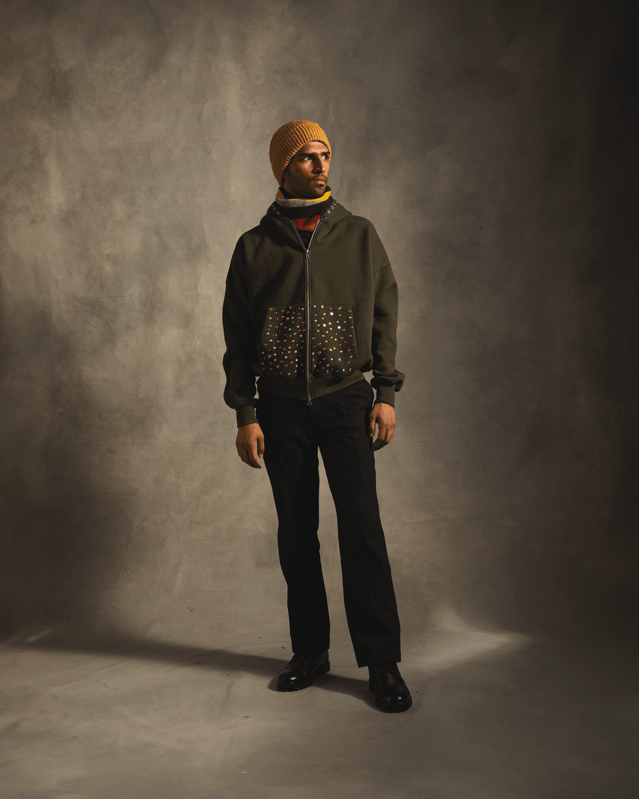Man in olive green zip-up hoodie with multicolored polka dot pockets, black pants, yellow knit beanie, and black shoes standing in studio